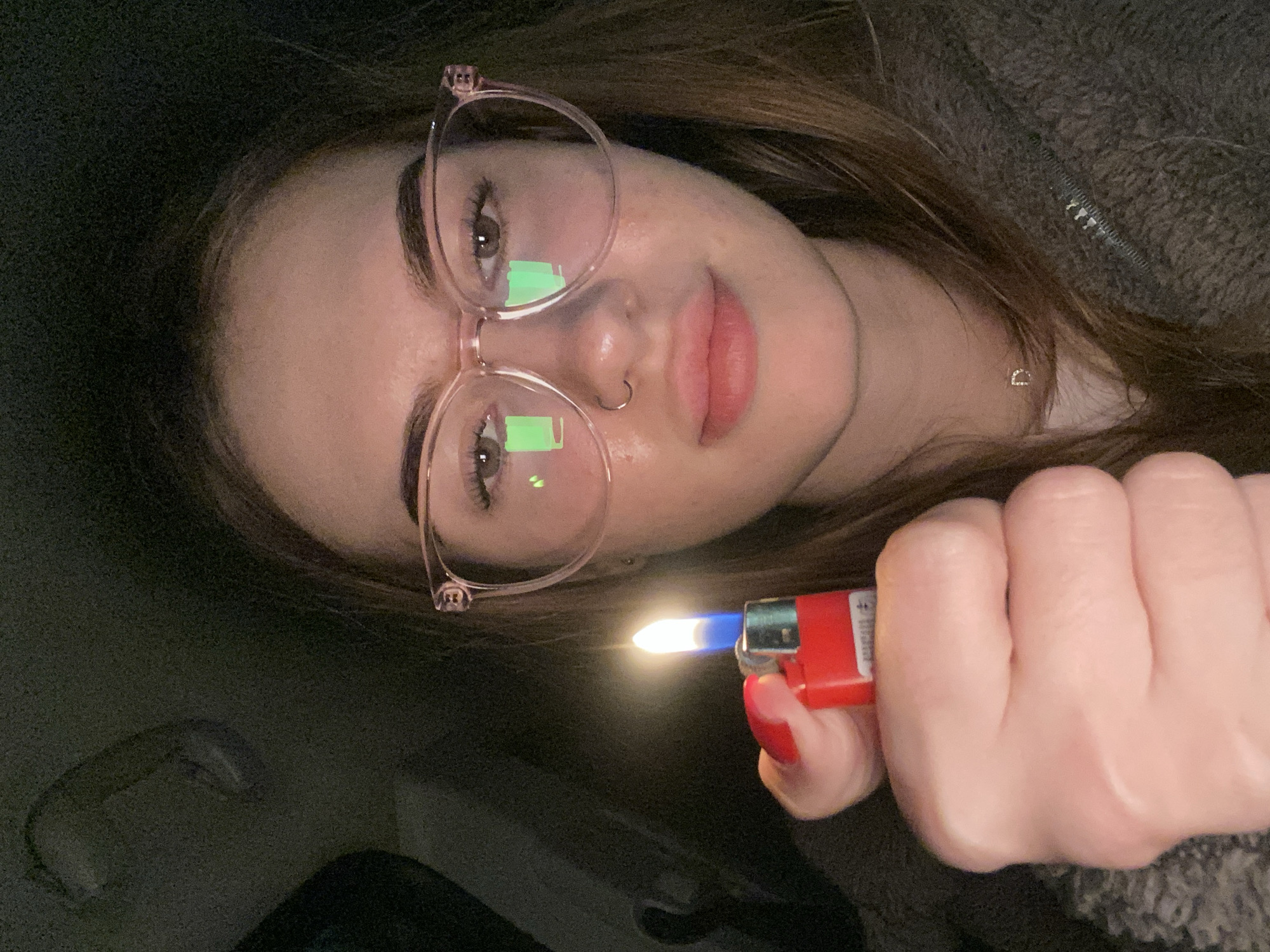 A close-up, horizontal portrait captures a young individual, likely female, in a low-light, possibly indoor setting. She is seen from the chest up, with her head tilted to the right, looking directly towards the camera.
The primary subject has light skin, long brown hair, and wears large, light pink or clear-framed glasses. Rectangular, greenish light reflections are prominent on the lenses of her glasses, reflecting into her eyes. She has dark eyebrows and a small hoop piercing in her left nostril. Her lips are full with a natural, rosy hue.
She is wearing a dark, possibly fleece or textured fabric, top or jacket, visible around her shoulders and neck. A small, subtle, possibly heart-shaped, silver or white necklace is visible near her collarbone.
In her right hand, held in a fist, she grasps a red disposable lighter with a bright blue and yellow flame emanating from it. Her fingernails are painted with red polish.
The background is uniformly dark and appears grainy, suggesting a dimly lit room or car interior. The overall impression is a casual, intimate portrait taken in low light. No legible text is visible on the lighter or elsewhere.
