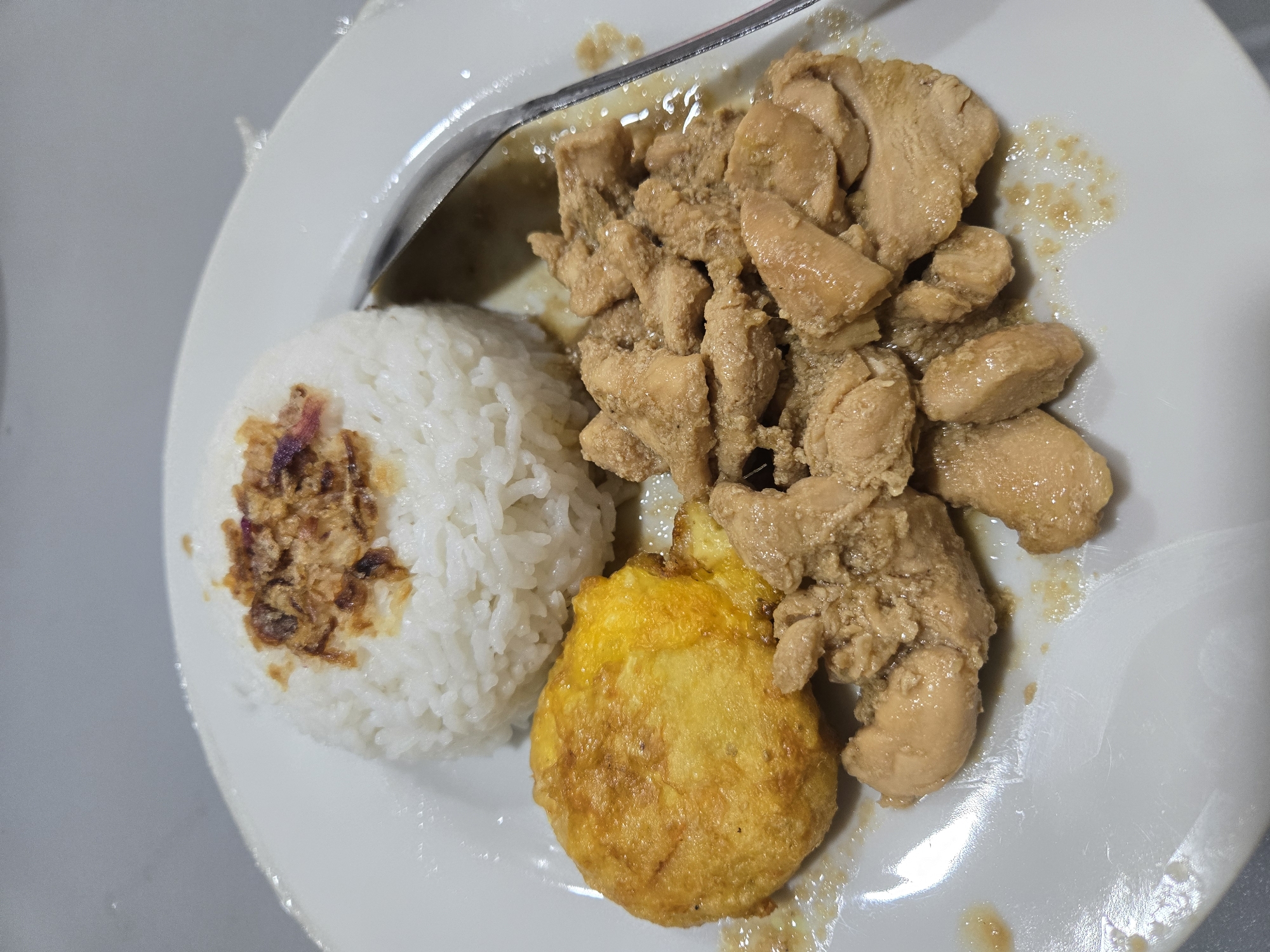 A close-up, overhead shot features a white plate with a meal. The meal consists of a mound of white rice topped with fried shallots, a fried egg, and a generous portion of sliced chicken in a savory sauce. A silver spoon rests on the right side of the plate, partially submerged in the sauce. The plate is placed on a gray, textured surface. There are no people visible in the image. The scene is indoors, likely in a kitchen or dining area. The image is brightly lit, with some glare visible on the plate and sauce. The meal appears to be Indonesian cuisine, possibly "Ayam Bumbu Rujak" or a similar dish, and the location is stated to be Sungailiat, Indonesia.