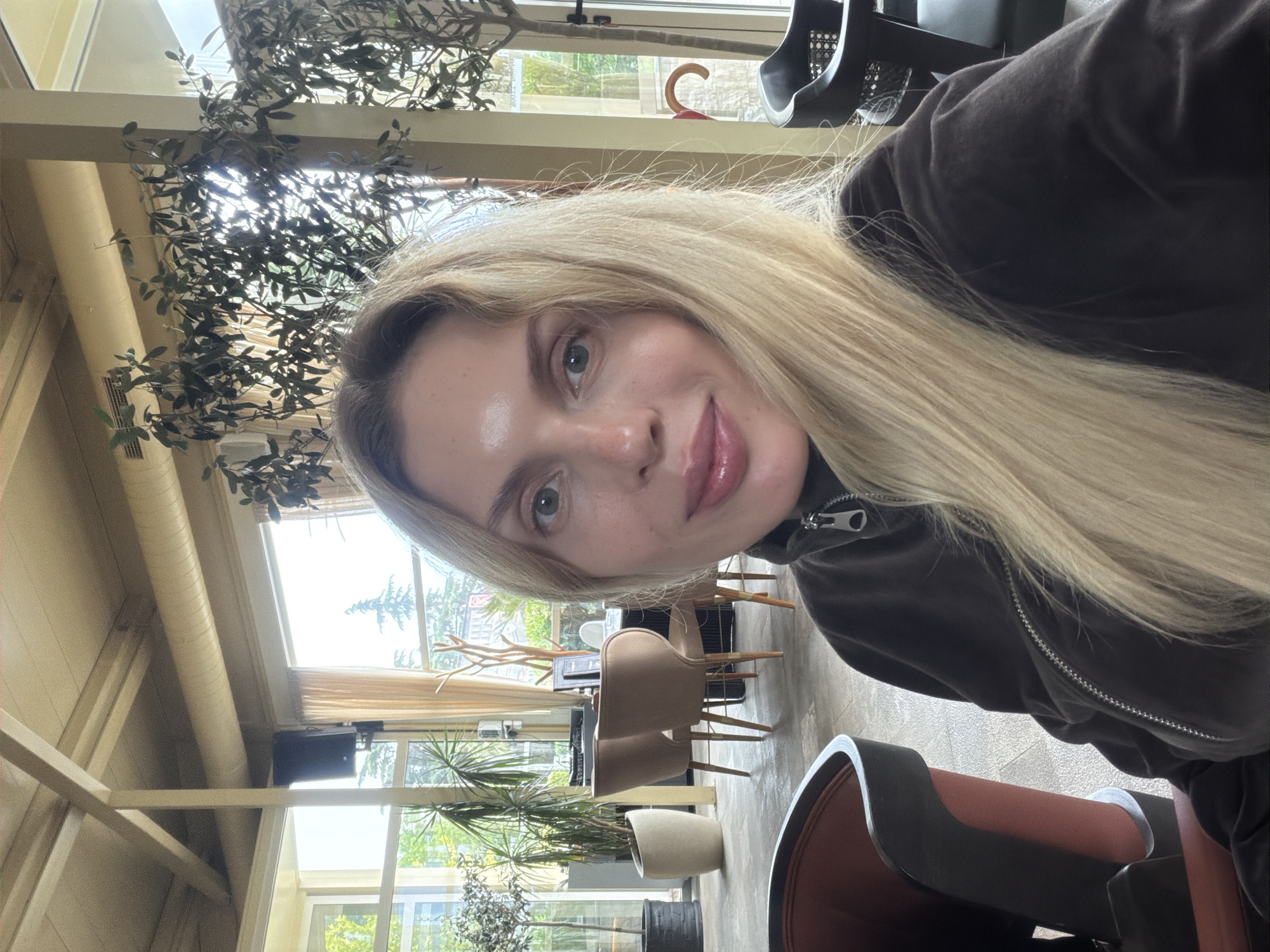 The image is a selfie taken indoors, likely in a café or restaurant with a greenhouse aesthetic. The subject is a blonde woman with light skin and visible freckles. She is wearing a dark brown jacket with a zipper pulled up to her neck.
The background features numerous windows providing ample natural light, revealing a view of greenery and what appears to be an urban setting. The indoor space has plants of various sizes, suggesting an attempt to integrate natural elements into the interior design. There are tables and chairs visible, some empty and some potentially occupied.
Based on the visible architecture, landscaping, and overall ambiance, it is plausible this location is in Budva, Montenegro, given its prevalence of urban greenery and tourist-oriented establishments.