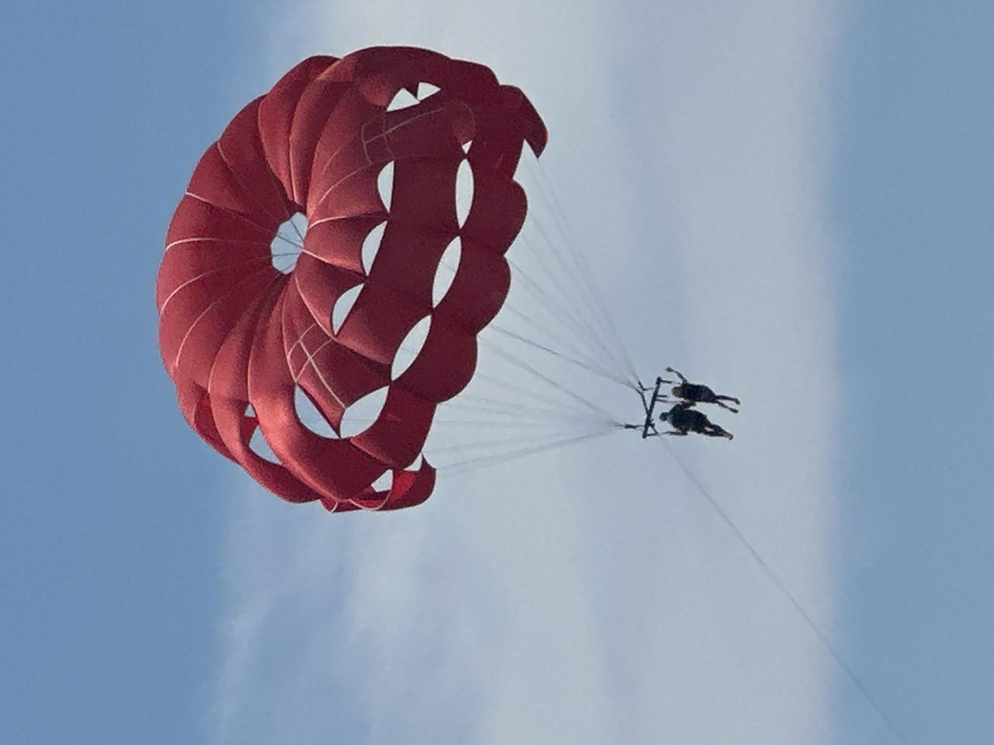 A large, bright red parasail is suspended in a bright blue sky with scattered white clouds. Two people are visible, suspended beneath the parasail by ropes and a harness. They appear to be enjoying a recreational activity, possibly parasailing over the water. The scene is serene and visually striking, with the vibrant red of the parasail contrasting against the pale blue of the sky and the white of the clouds.