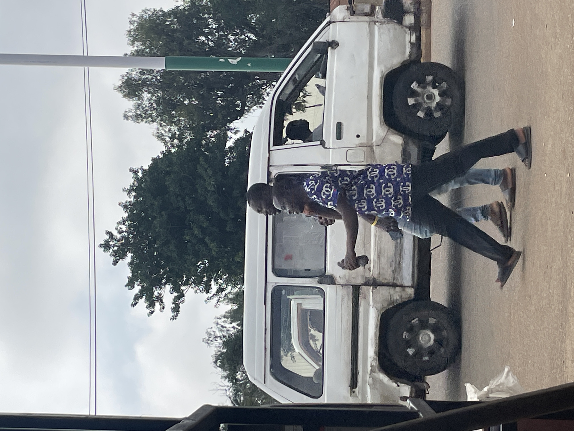 The image shows a street scene in Bukuru, Nigeria. Two men are walking in front of a white van. The man in the foreground is wearing a blue shirt with a white pattern and black pants. He is holding a dark object in his right hand. The man behind him is partially obscured, wearing a blue shirt. The white van appears to be old and somewhat worn. The van's doors are open, and a person can be seen sitting in the driver's seat. Trees are visible in the background, along with a utility pole. The ground is paved, and the sky is overcast. The overall impression is a candid snapshot of everyday life on a Nigerian street.