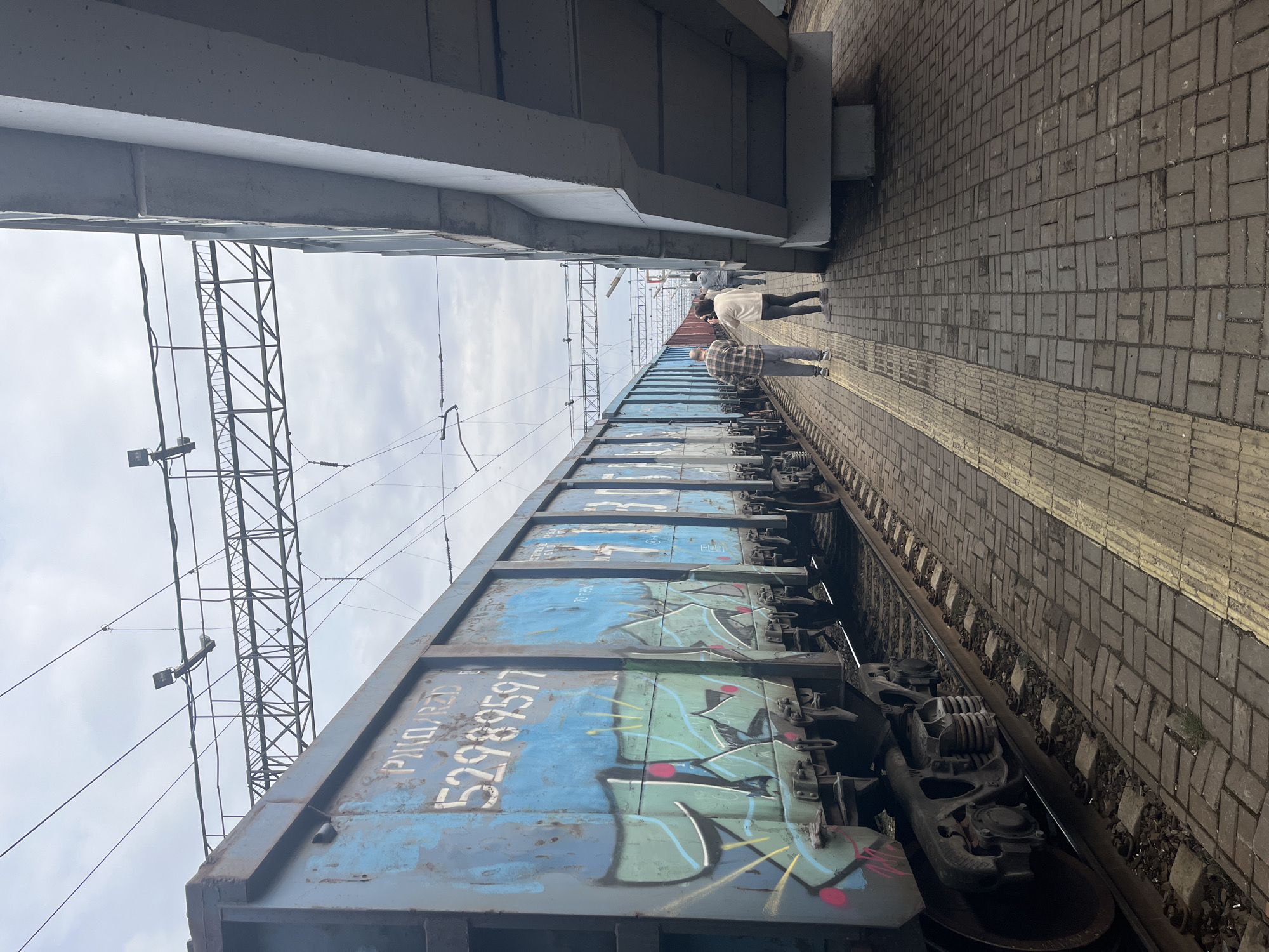 The image depicts a railway platform in Agryz, Russia. A blue freight car with the number "52989597" marked on its side, covered in graffiti, sits on the tracks. Overhead electrical wires and a support structure are visible. People are walking along the platform beside the train. A concrete bridge or overpass is above the platform. The ground is paved with rectangular paving stones. The sky is overcast.