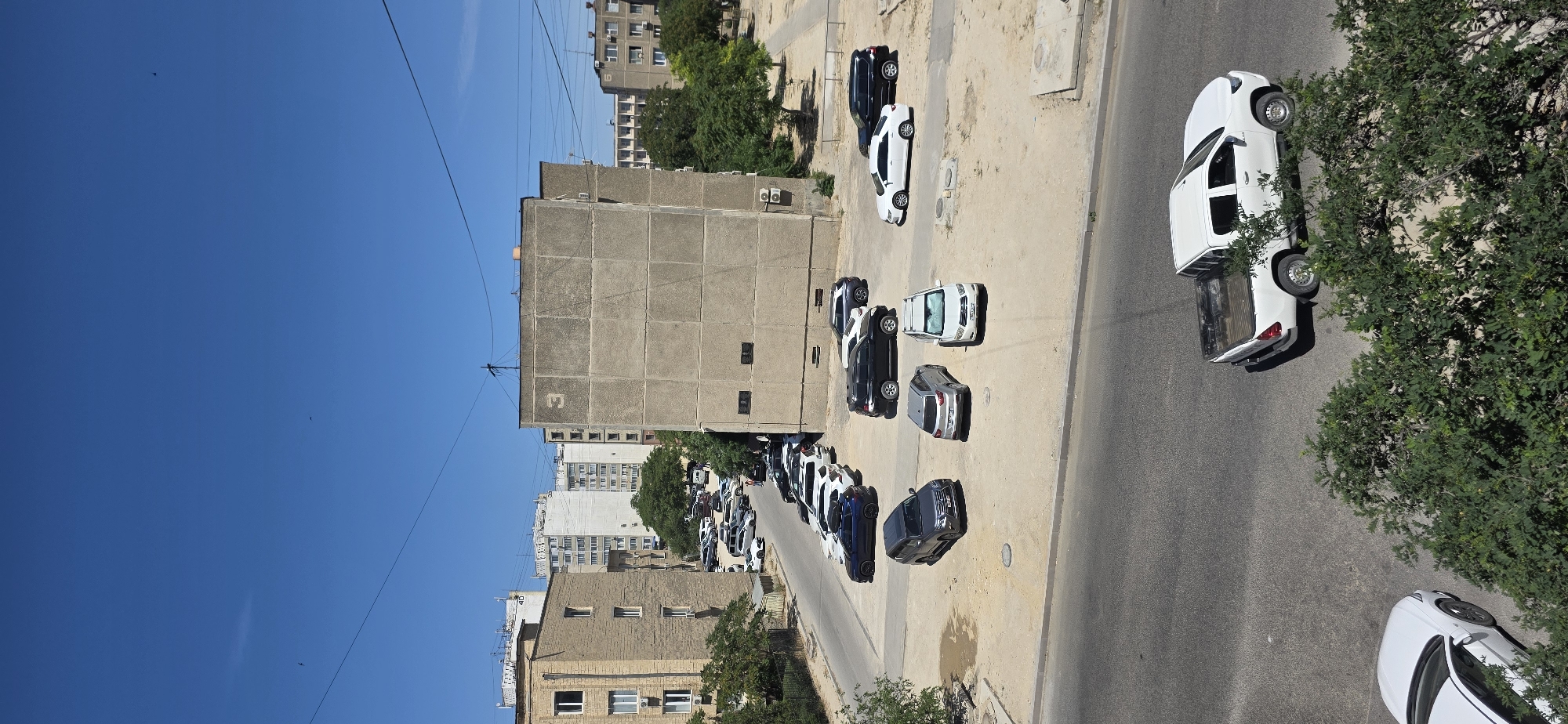 This is an aerial, vertical-oriented image of an urban scene in Aktau, Kazakhstan. The sky is a clear, vibrant blue, suggesting a sunny day. The scene is dominated by multi-story residential buildings and a street lined with parked cars. The architecture appears to be Soviet-era, with concrete facades and balconies.
The primary subjects are the numerous vehicles, mostly cars and SUVs, parked along the road and on a dirt lot adjacent to the buildings. They are of various colors including white, black, and silver. A white pickup truck is visible parked near some dense green foliage. The buildings themselves are also prominent, with visible details like air conditioning units mounted on some of them and windows. Some of the buildings have numbers visible, such as a "3" on one concrete structure.
The activity taking place is the ordinary urban life of residents, indicated by the parked cars suggesting people are home or nearby. There is no visible activity of people in motion. The time of day is clearly daytime, given the bright sunlight casting shadows. The weather appears to be good, with a clear sky.
Notable details include the power lines crisscrossing the sky and the slightly uneven, dusty ground in the parking areas. The overall impression is of a typical residential neighborhood in a city, possibly a bit aged, under a clear, bright sky.