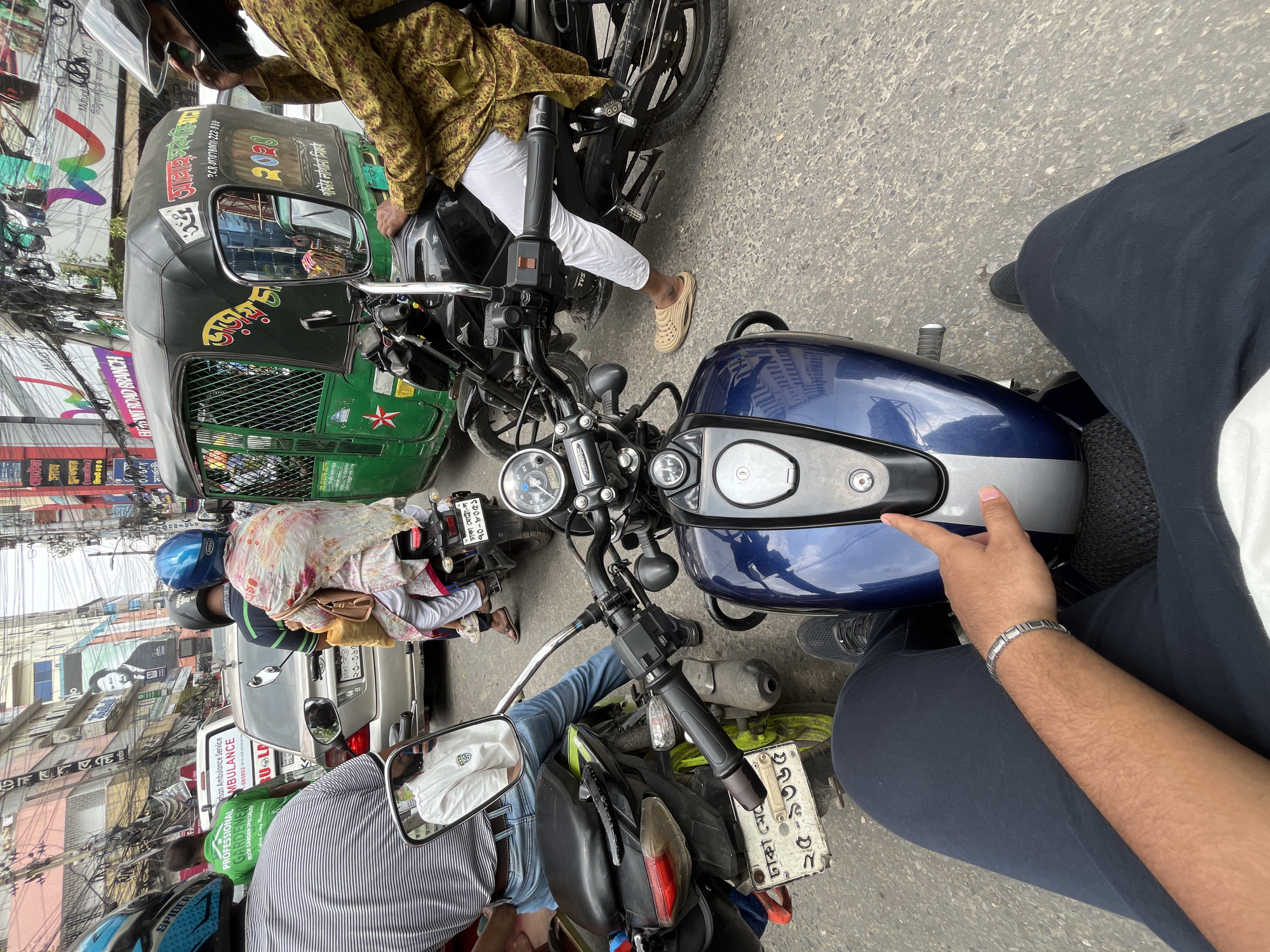 The image captures a scene in Dhaka, Bangladesh, likely during daytime street traffic. The view is from the perspective of someone sitting on a blue motorcycle. Other vehicles are nearby, including a green auto-rickshaw ('CNG' or 'Baby Taxi') with the registration number "ঢাকা মেট্রো -ঠ ১১-৯৮৫৮", and other motorcycles. A person wearing a patterned tunic and white pants sits on a motorcycle beside the blue one. A number plate is visible on one motorcycle reading "ঢাকা মেট্রো-ল ২৫-৯৩৬৫". Buildings with numerous signs and electric wires overhead line the street. The overall impression is of a congested urban environment.