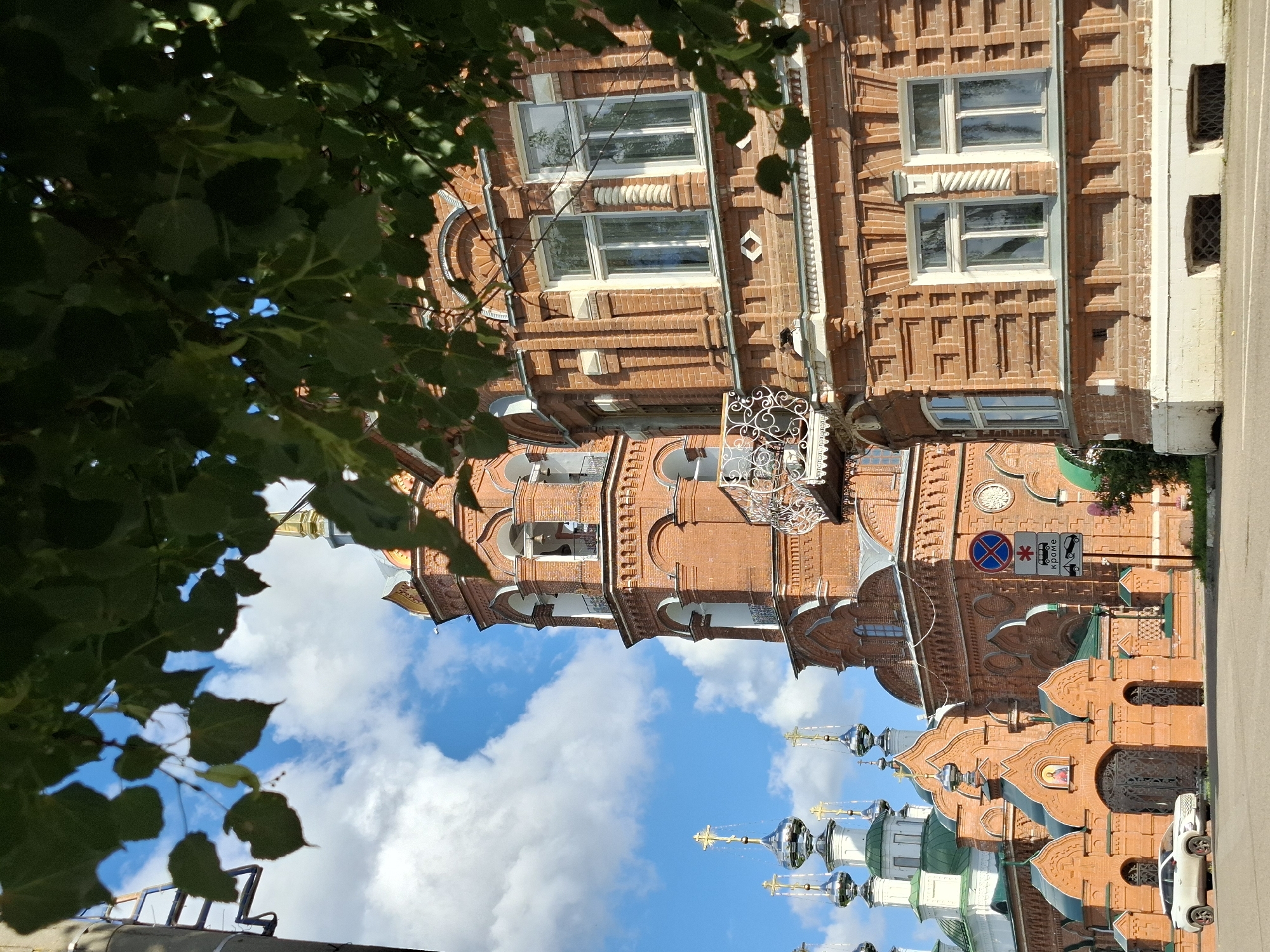 The image shows a street view of the Assumption Cathedral in Kungur, Russia. The cathedral, constructed of red brick, features intricate architectural details, including multiple domes topped with crosses, a prominent bell tower, and decorative window surrounds. The right side of the image is dominated by a large, multi-story brick building with white-framed windows. Green foliage from trees partially obscures the view of the sky and parts of the buildings. A white car is parked on the street in front of the cathedral's entrance. Traffic signs are visible near the cathedral. The sky is blue with white clouds, suggesting it is daytime.