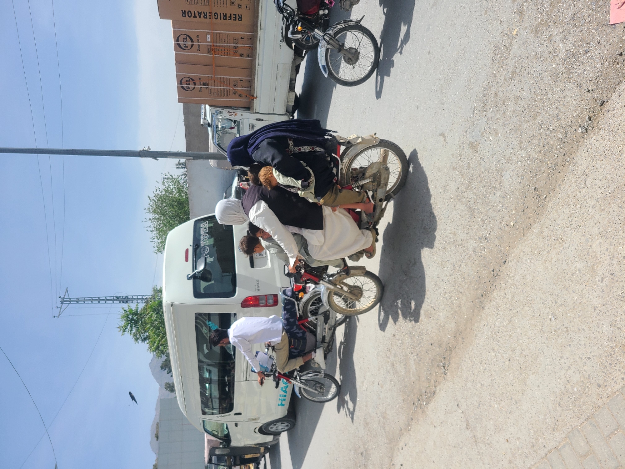 The image depicts a street scene in Quetta City Tehsil, Pakistan, with multiple motorbikes and vehicles present. Several individuals are riding motorbikes; on one bike, at least three people are seen, one wearing a hijab. Another motorbike is carrying goods, appearing to be stacked boxes of refrigerators. A white passenger van is present. The location appears to be an urban area with paved roads and overhead power lines. The sky is clear and bright, suggesting it is daytime.