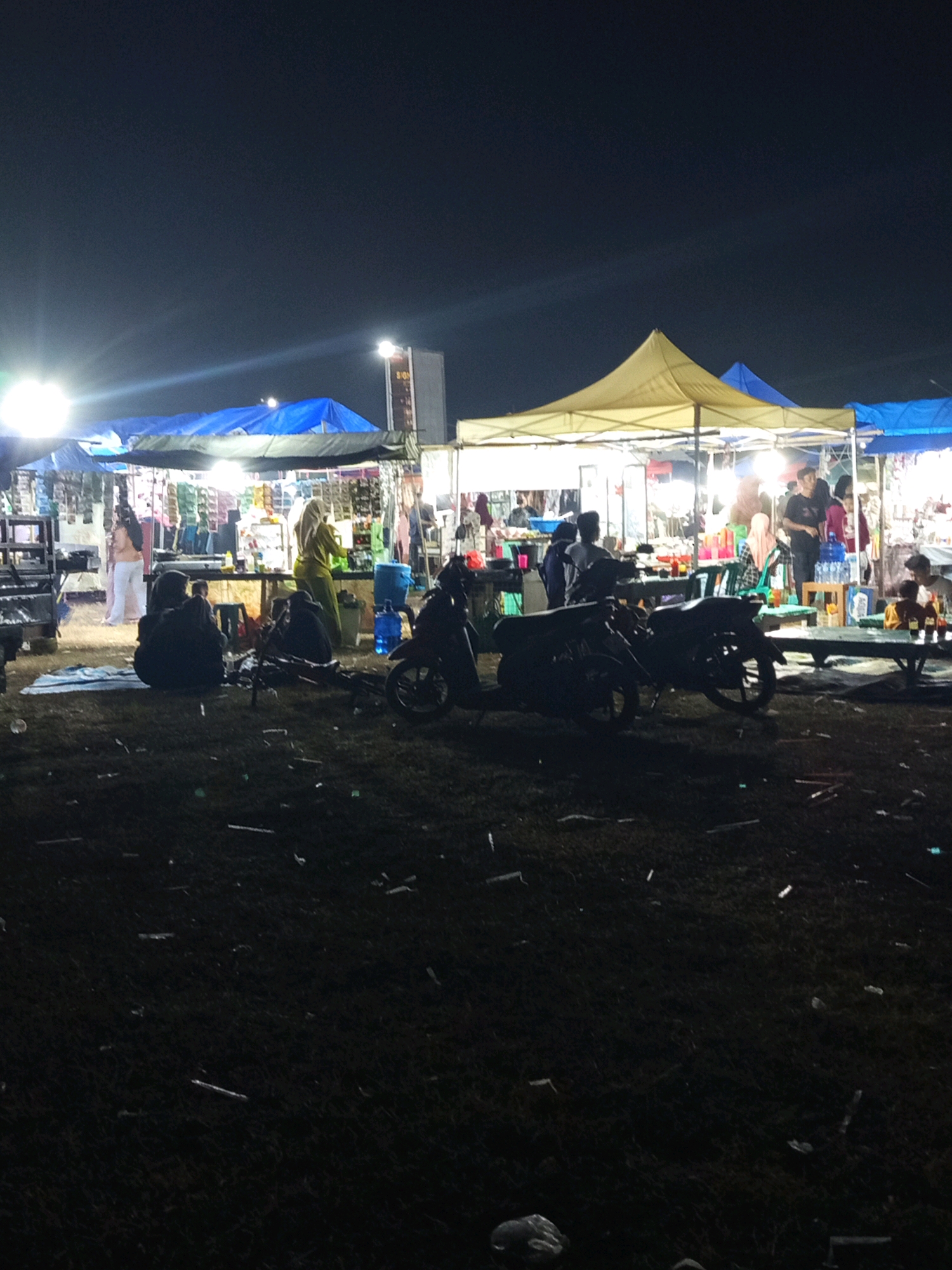 The image depicts a night scene at an outdoor market in Pulung Kencana, Indonesia. In the foreground, grass covers the ground with scattered debris. Several motorcycles are parked, silhouetted against the dimly lit environment. In the mid-ground, a market is set up with various stalls covered by tents. Yellow and blue tents are visible. People are seen browsing and interacting within the market stalls. One stall has items displayed on shelves, and another shows goods on a table. A blue water dispenser is positioned near a stall. In the background, a tall, illuminated building with a sign reading "SION" is visible. The overall atmosphere is that of a lively night market.