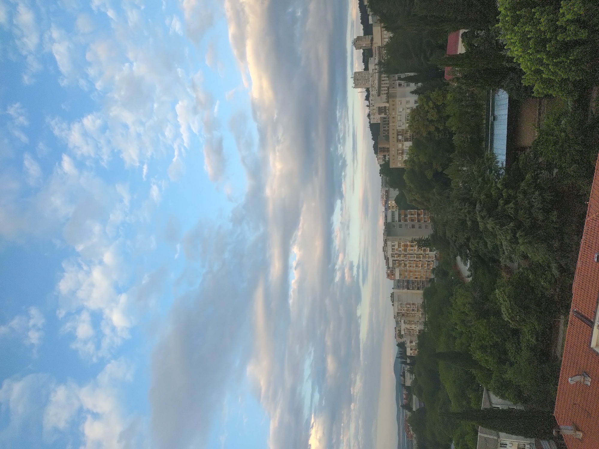 The image shows a panoramic view of a city skyline in Stara Zagora, Bulgaria, under a partially cloudy sky. The foreground features a red-tiled roof and some green trees. In the midground, several apartment buildings of varying heights are visible, interspersed with lush green trees. Some of the buildings appear to be older Soviet-era blocks, while others are more modern. The background shows a hilly landscape with more buildings and a few taller structures, possibly communication towers. The overall scene suggests a residential area within the city, with a mix of architecture and abundant greenery. The lighting indicates it is either early morning or late afternoon, with soft, diffused light. No people or specific actions are discernible in the image.