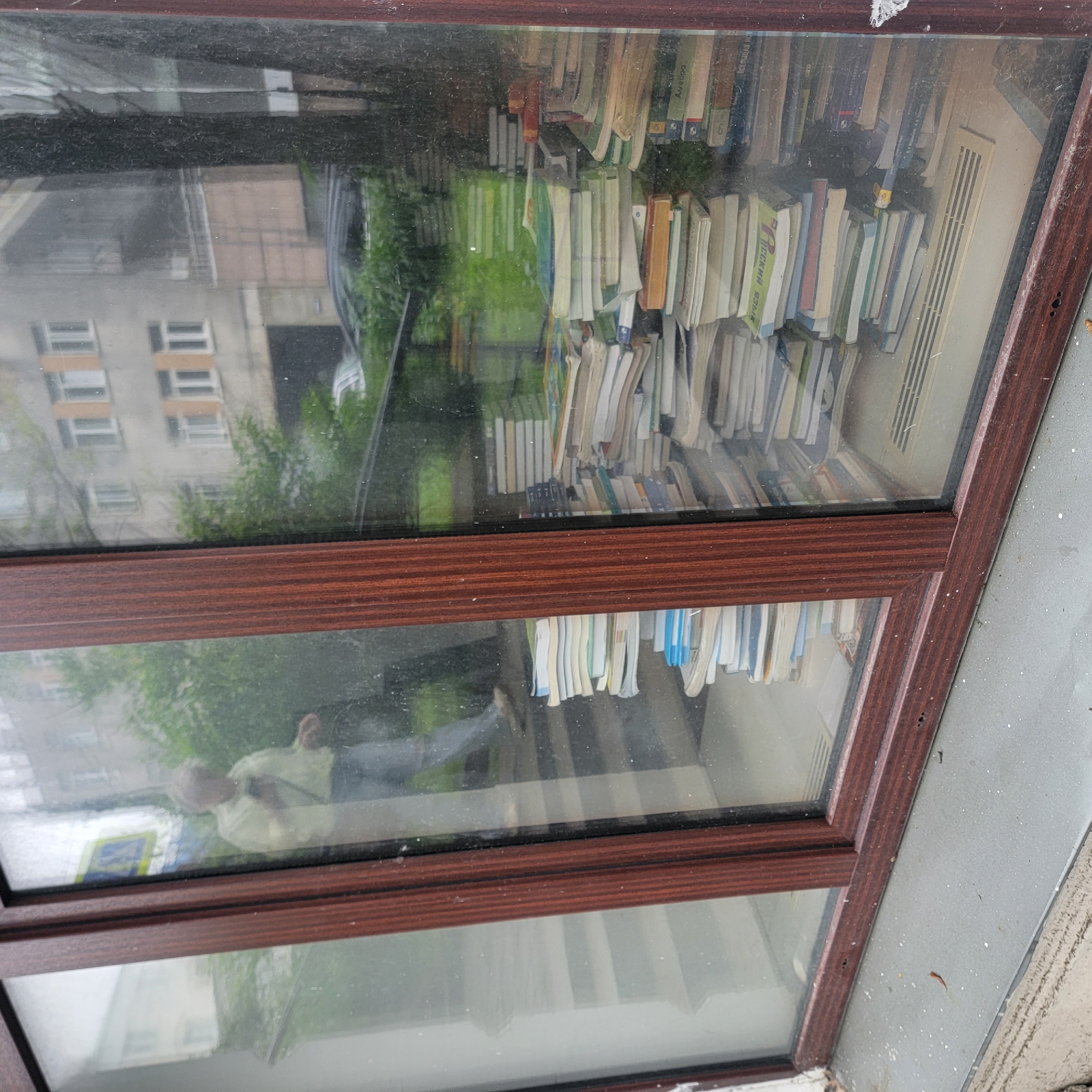 A person is reflected in a dirty window with a wooden frame. The window overlooks a street scene in Vladivostok, Russia. A building with multiple windows and balconies is visible across the street, along with trees and parked cars. Inside the window, stacks of books are piled up, some on shelves and others on the floor. The books appear to be old and well-read. The person in the reflection is wearing a light-colored shirt and jeans and is holding a phone. The overall impression is of a slightly cluttered and lived-in space, possibly a home or a small shop.