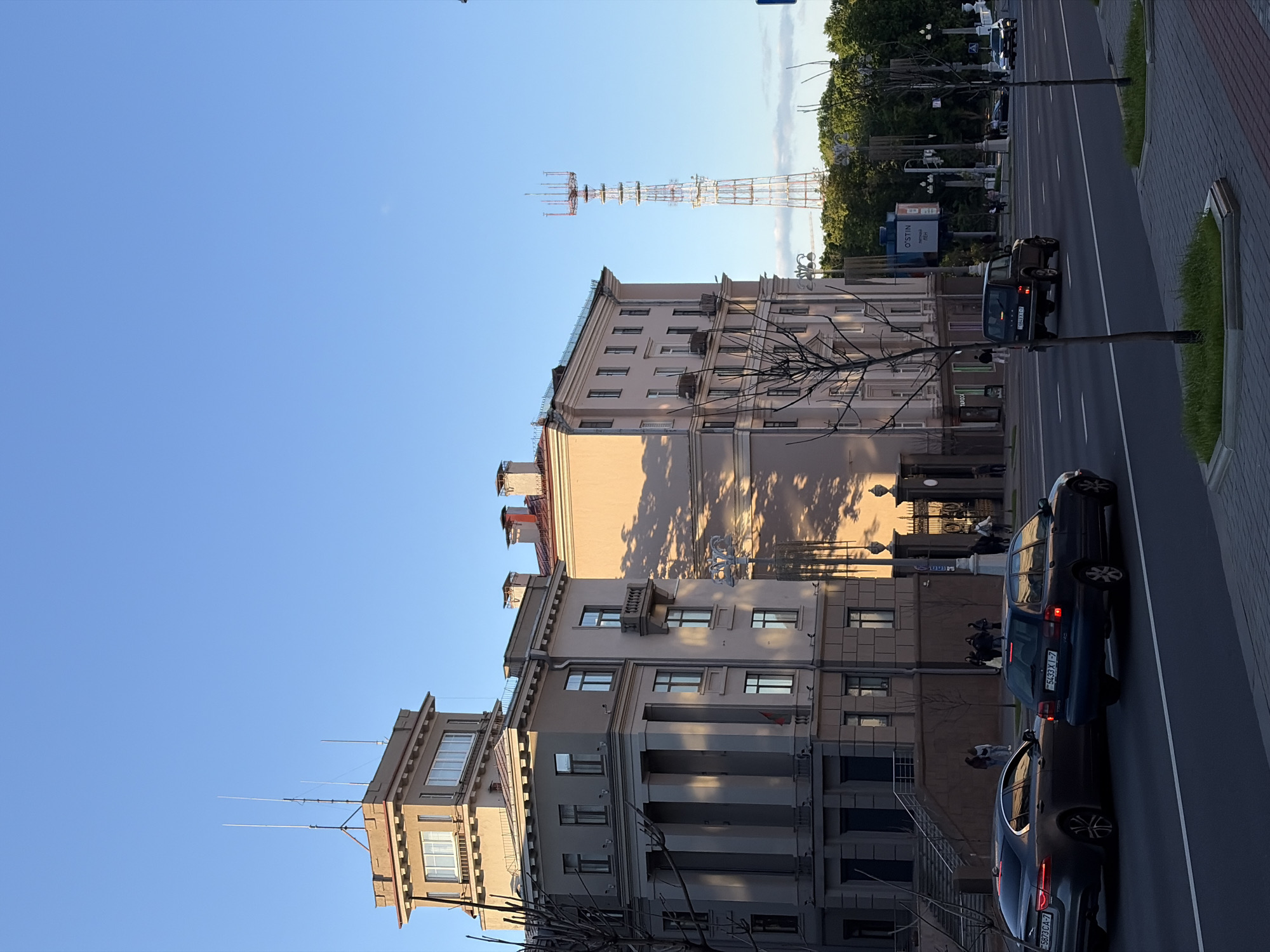 The image shows a street scene in Minsk, Belarus. Buildings line the left side of the road, displaying multi-story architectural styles with columns and varying window arrangements. A communications tower is visible in the background. Several cars are parked along the roadside. Bare tree branches appear in the foreground, casting shadows on the buildings. People are seen walking along the sidewalk. The sky is a clear blue. The street is divided by painted white lines. Vegetation is on the right edge of the image.