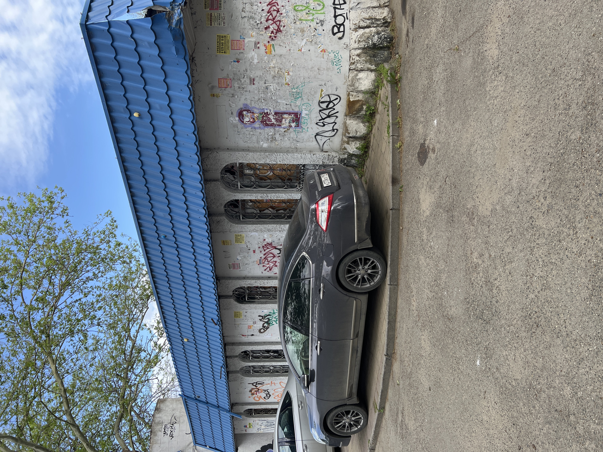 The image depicts a gray Nissan Teana parked on a street next to a building. The building has a blue corrugated metal roof and a gray wall covered in graffiti and posters. There are four arched windows on the wall. The car's license plate number is partially visible as X273XX 193. Telephone numbers and Cyrillic text are visible on posters. The environment appears urban with a mix of graffiti, architecture, and vegetation. Based on the vehicle's license plate and the Cyrillic text, this is probably located in Russia. The phone numbers seem Russian. Taking all of these into account, it is likely Krasnodar, Russia.