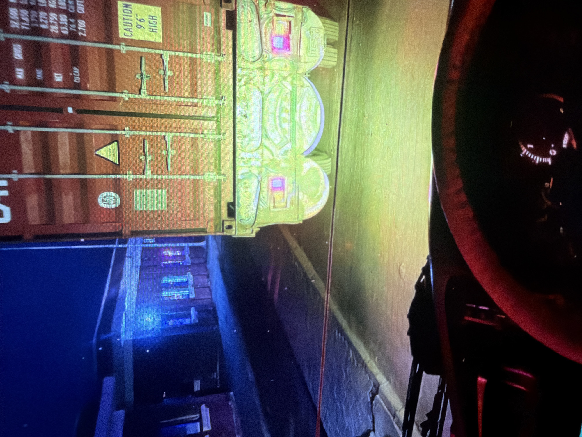 The image depicts a nighttime view from the interior of a truck cab, looking out towards a shipping container at what appears to be a port or industrial area. The truck's dashboard and steering wheel are partially visible in the foreground, with a red steering wheel cover and illuminated gauges. The main subject of the scene is a large, red shipping container, seen from the rear, with various markings and labels. One prominent label reads "CAUTION 9'6" HIGH". The background shows a dimly lit building with blue and pink lights, suggesting it could be a control room or office. The ground is paved and there are visible tire tracks. The overall atmosphere is dark, with artificial lighting creating distinct shadows and highlights. The location in Bukuru, Nigeria, is not explicitly visible within the image itself, but is provided as context.