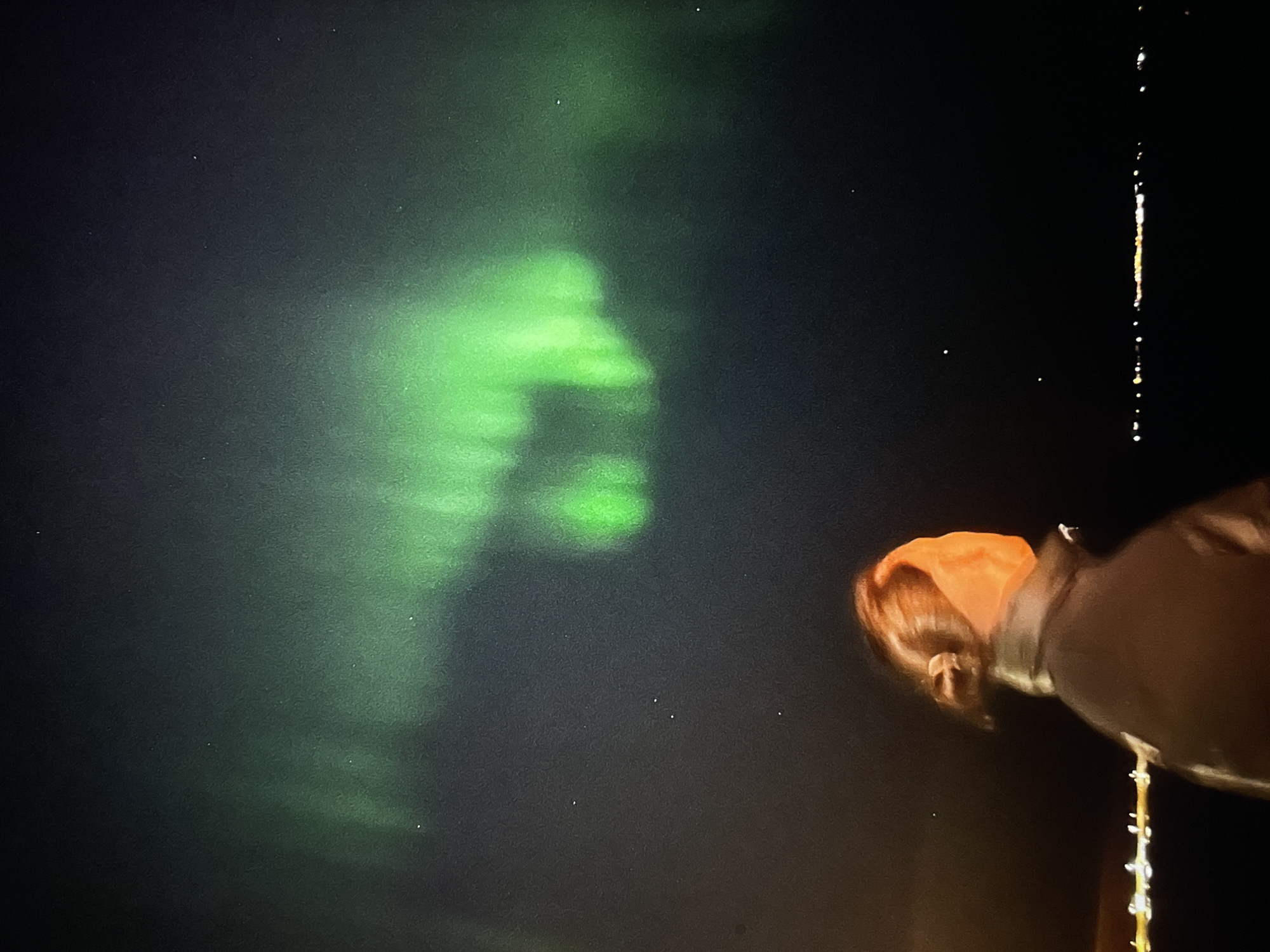 The image depicts a night scene with an aurora borealis dominating the left side of the frame. The aurora exhibits a diffused, green glow. On the right side of the frame, there's a person wearing a dark jacket with an orange/red head covering. The location appears to be outdoors, given the aurora and the lack of visible structures. It is impossible to determine the location in the city of Unknown, Unknown, based solely on this visual information.