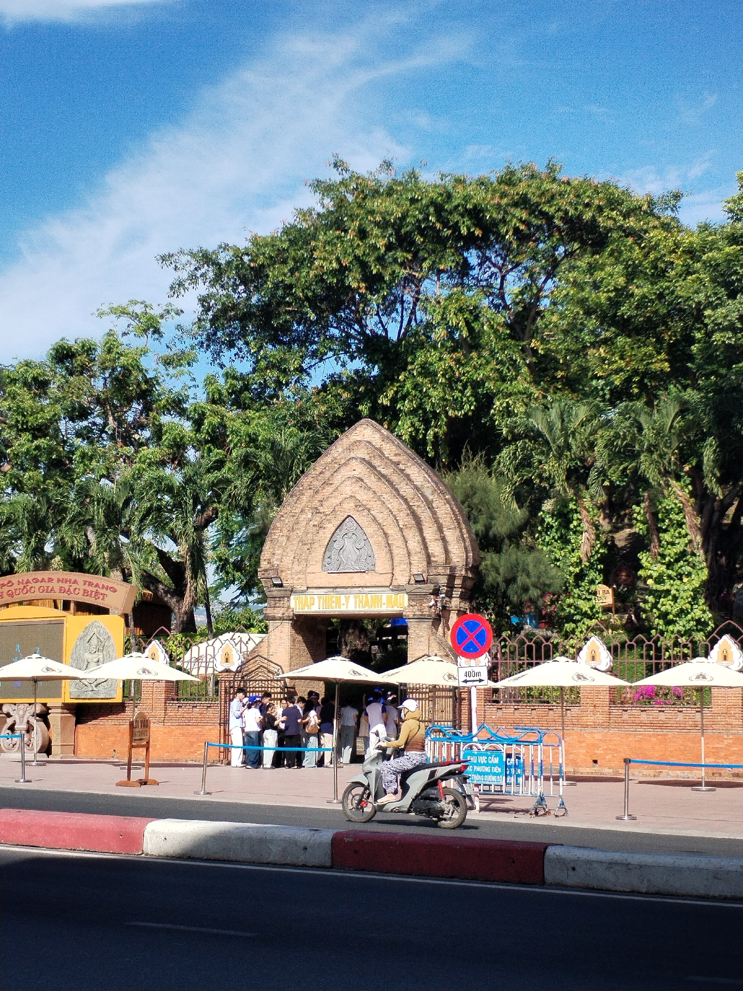 The image depicts an outdoor scene in the city of Bac Nha Trang Ward, Vietnam, likely at or near a historical site. In the foreground is a paved street with a red and white curb, behind which a pedestrian walkway is located. A person is riding a scooter along the sidewalk. Further back is a brick fence with decorative white lotus-shaped elements along its top edge, with several white umbrellas providing shade over the walkway. Behind the fence, a group of people appears to be gathered near the entrance of a brick structure with an arched top. The structure is surrounded by trees and greenery. A blue and red prohibition sign is visible near the entrance, with the text "400m" on it. To the left is a signpost with text in Vietnamese, with the words "PO NAGAR NHA TRANG" visible. The sky above is blue with some clouds.
