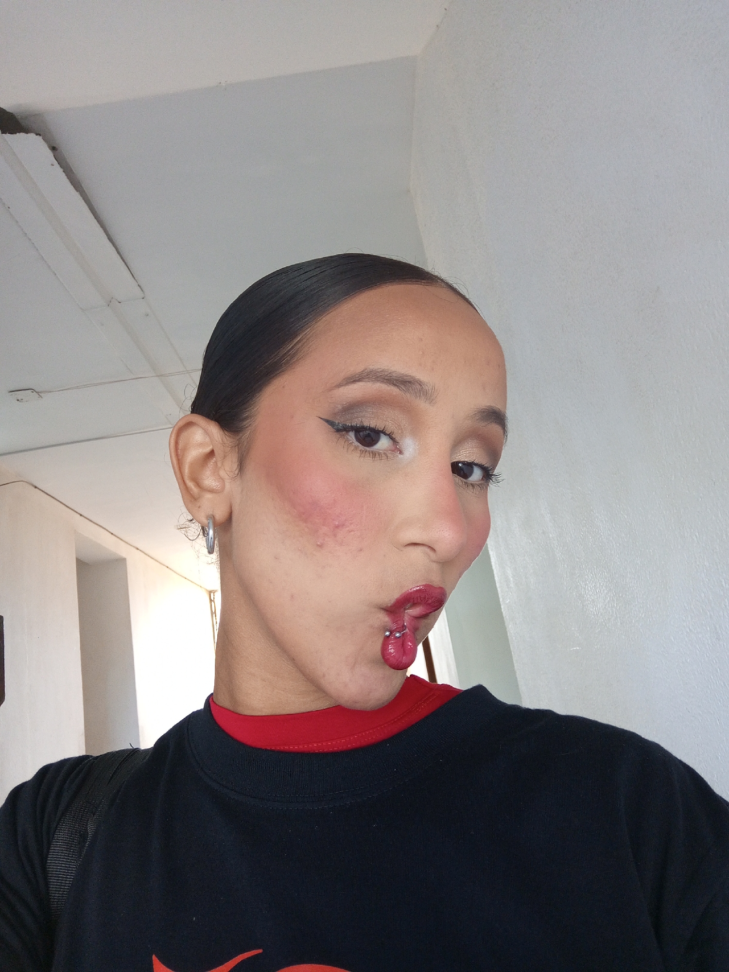A person with dark hair pulled back into a sleek style, wearing makeup including defined eyebrows, eyeshadow, eyeliner, blush, and glossy red lipstick, is posing indoors with a slightly distorted facial expression. The person has a tongue piercing visible. They are wearing a black long-sleeved shirt with a red collar peeking out from underneath. The person is wearing silver hoop earrings and appears to have a black backpack slung over their shoulder. The background consists of a white wall and a white ceiling with a small portion of what appears to be a hallway visible. The location is not discernible from the image.