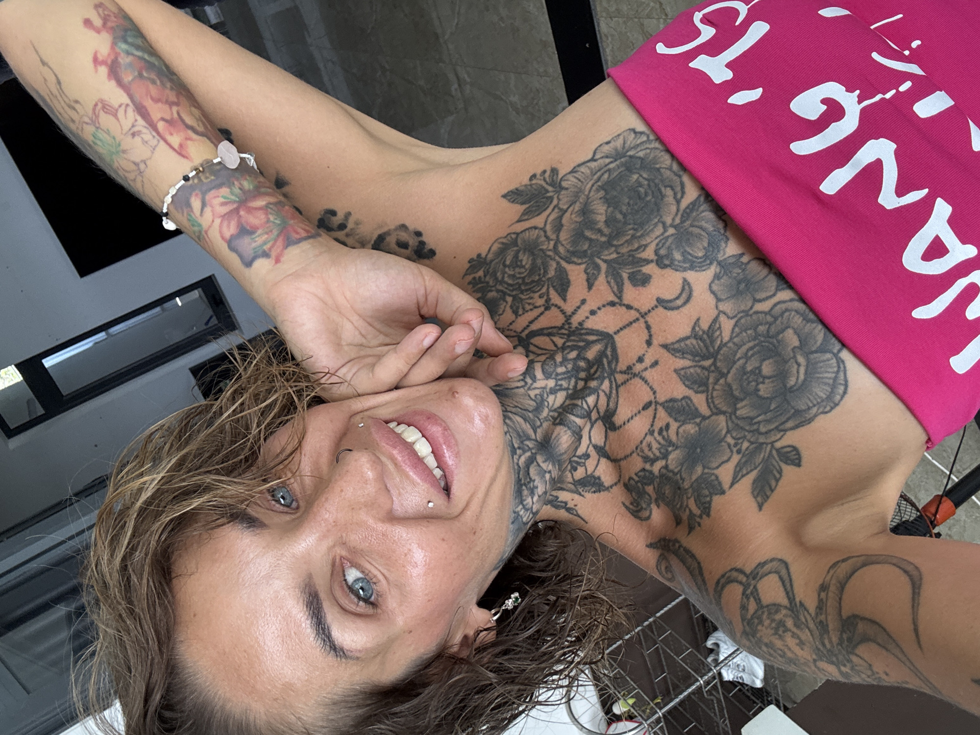 A woman with wet, short brown hair and blue eyes is posing for a selfie. She is smiling and has several piercings, including a nose ring and two lip piercings. She is heavily tattooed on her chest, neck, and arm, with floral and other designs. She is wearing a bright pink strapless top with white text. The background appears to be an indoor setting with natural light, possibly a hotel room or apartment, with a glimpse of outdoor scenery through a window or door. The provided location of Bac Nha Trang Ward, Vietnam, is not discernible from the visual information within the image.
