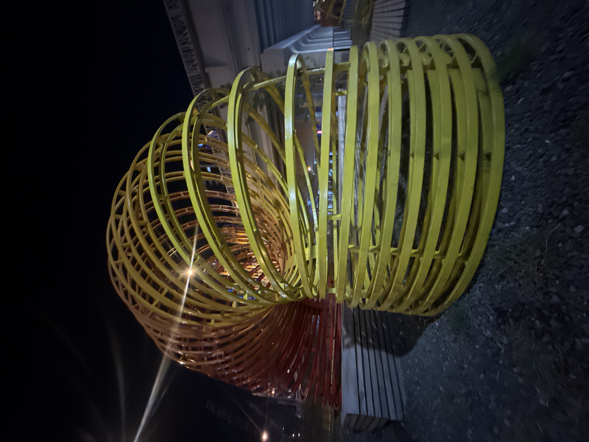 The image depicts a large, spiraling metal sculpture, likely an art installation, located outdoors at night. The sculpture consists of a series of concentric rings, painted in a gradient from red/orange at one end to yellow at the other. It appears to be constructed from metal bars. The sculpture is positioned next to a dark, gravelly area and a wooden bench or platform. In the background, there are blurred lights, possibly from street lamps or buildings. The photograph may have been taken in Yekaterinburg, Russia, based on the user's prompt, but specific visual details confirming this location are absent.