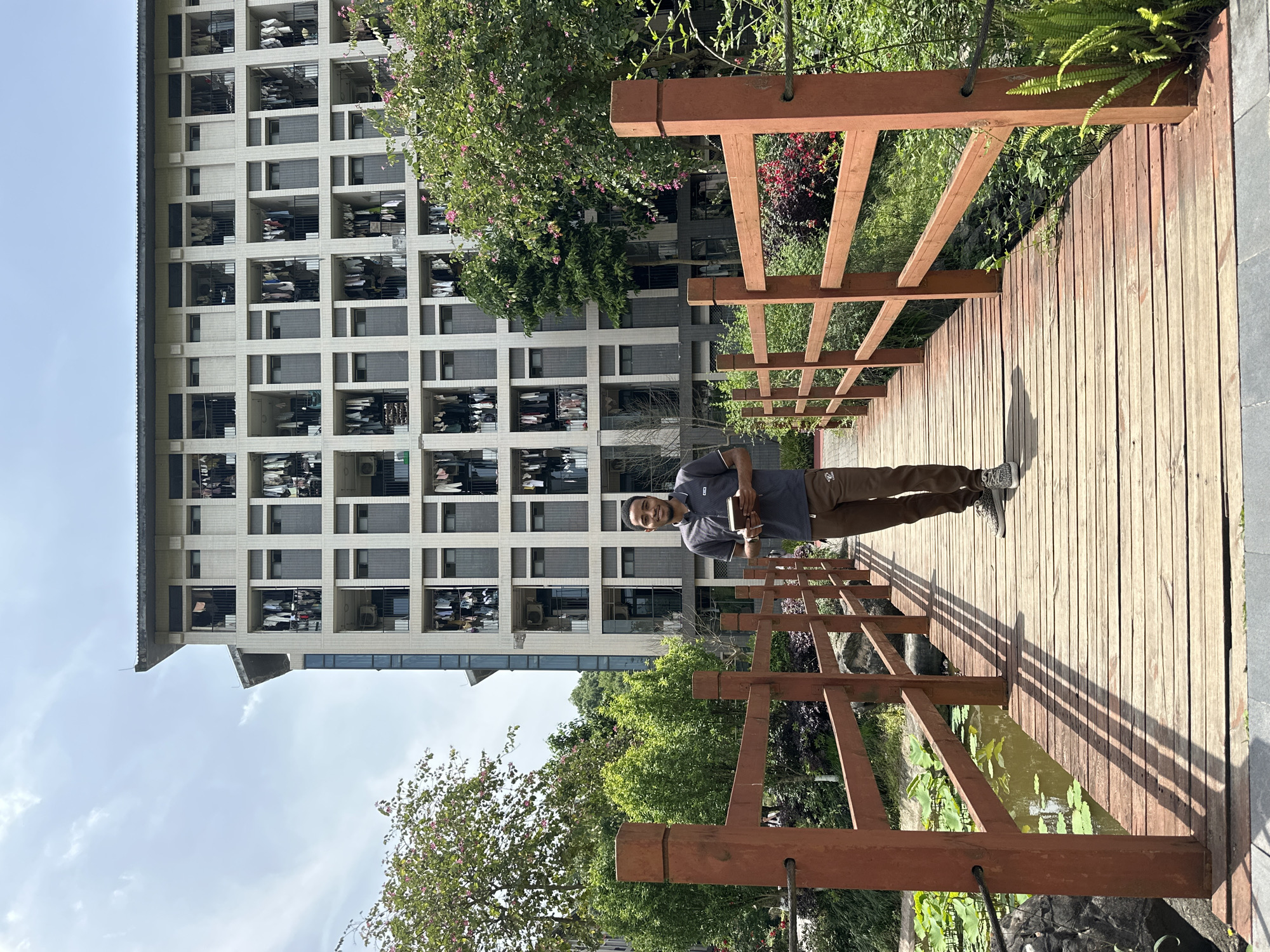 A man stands on a wooden bridge in Jiangjin, China. He faces the camera, holding a book. A tall building is visible in the background, its facade marked by numerous rectangular windows and balconies. The bridge spans over water with lily pads. Lush greenery surrounds the bridge, and a cloudy sky is overhead.
