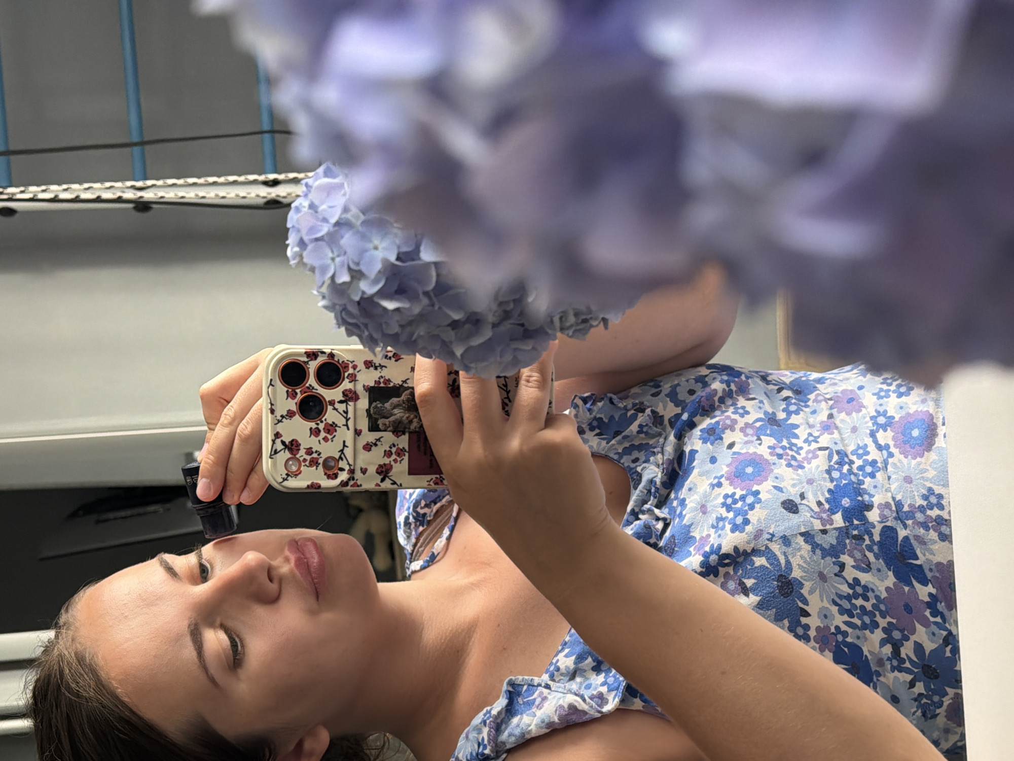 A young woman is captured in a close-up, waist-up reflection, likely in a home environment. She is holding a smartphone in her right hand and a small dark-colored object, possibly a cosmetic item, in her left hand, which she is bringing towards her face, seemingly applying it to her cheek. She is wearing a blue floral-patterned dress with ruffled straps. In the foreground, out of focus and to the right, is a large cluster of light purple hydrangeas. The background is softly lit, with hints of a mirror or reflective surface and what appears to be part of a window frame or blinds. There is no indication of the city of Sochi, Russia, in the image or its contents.