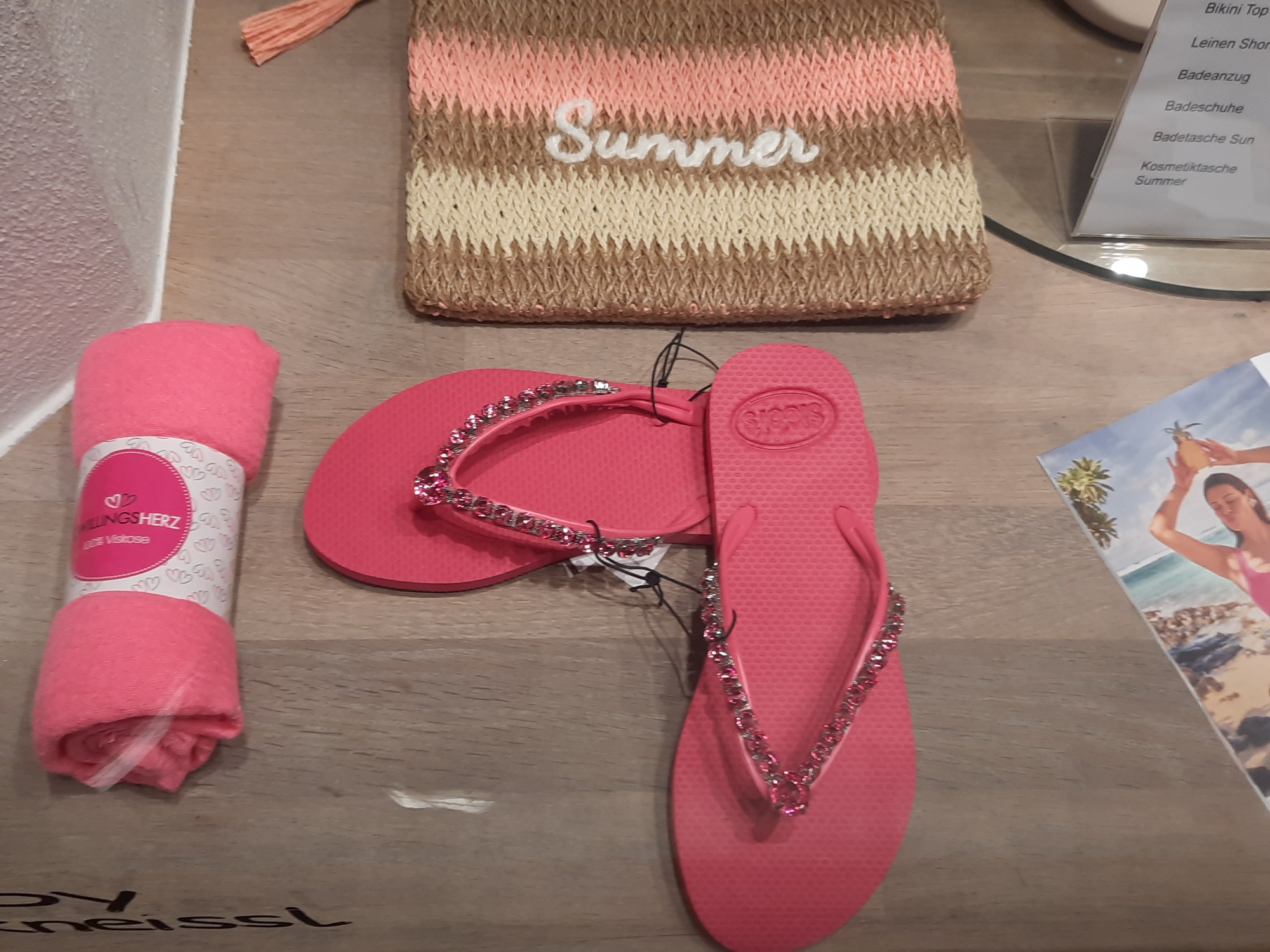The image shows a close-up of items laid out on a light-brown wooden surface. In the upper left, a rolled-up pink towel with a label is visible. To the right of the towel, a pair of pink flip-flops adorned with rhinestones is placed. To the right of the flip-flops is a small, woven bag with a striped pattern in beige, cream, and pink, and the word "Summer" is embroidered in white on its surface. A small portion of a brochure with an image of a beach scene and a woman is visible in the lower left corner. The background appears to be a wall with a textured surface. The overall impression is a display of summer-themed items, possibly in a retail setting. The items appear to be new and unused. There are no people visible in the image. The lighting suggests an indoor environment, possibly a shop. Without further context or identifiable landmarks within the image, it is not possible to determine the specific location in the city of Rosenheim, Germany.