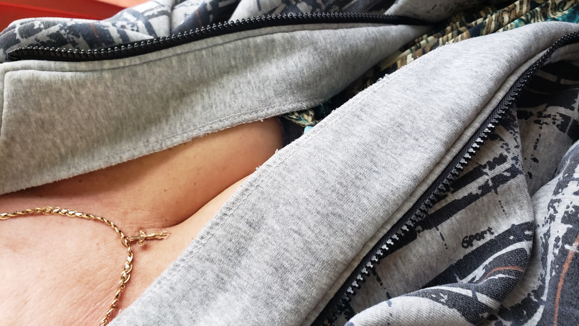 A close-up, eye-level shot focuses on the décolletage of a person wearing a gold chain with a crucifix pendant. The person is also wearing a grey hooded jacket with a zipper, revealing a glimpse of a patterned garment underneath. The background is out of focus and appears to be a red surface. The image contains no visible elements that would indicate a specific location within Podtureň, Slovakia.