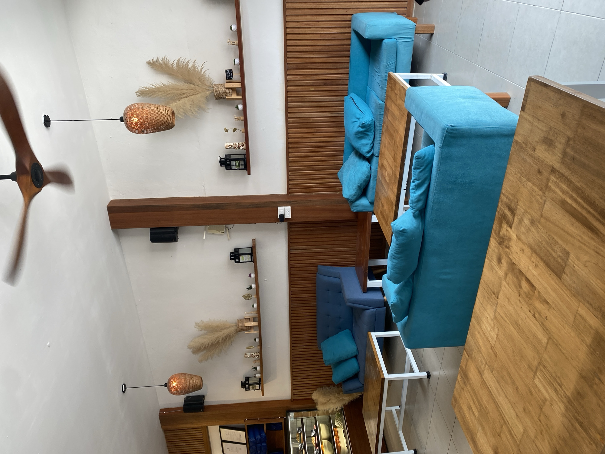 The image depicts an interior scene likely in a restaurant or cafe in Baan Chaloklum, Thailand. The space features linear seating arrangements with blue-upholstered benches accompanied by wooden tables. The tables have white metal frames. The benches and tables are positioned against a backdrop of wooden slat wall panels. Decorative elements include rattan pendant lamps, shelves adorned with pampas grass and small decorative items. A ceiling fan is visible in the upper left corner. Below the second seating area are glass coolers filled with various food. The room appears well-lit, likely from both natural light and the pendant lamps.