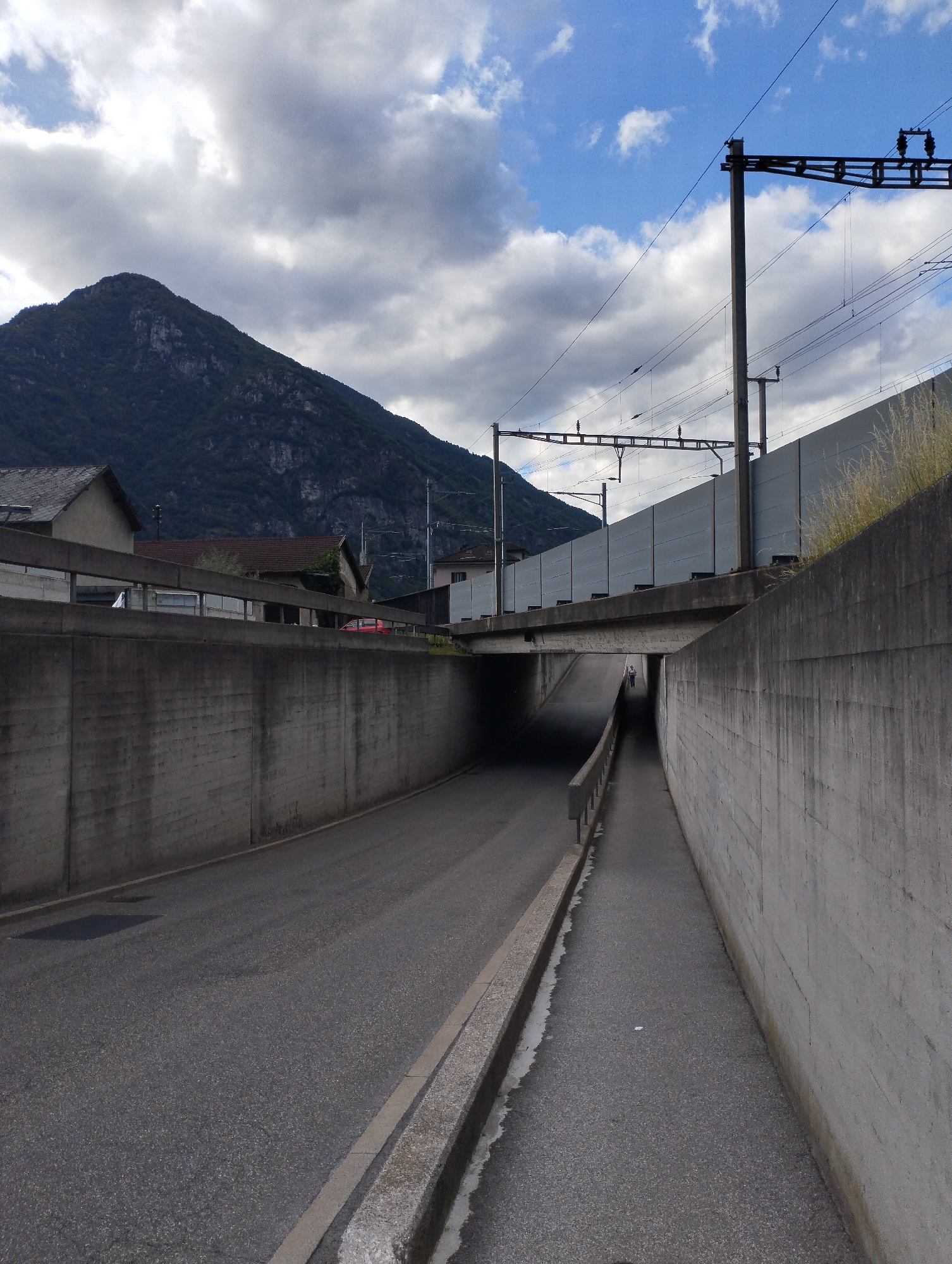 The image shows a view of a tunnel entrance and roadway in an urban environment. The roadway is paved and appears to be a two-lane road. There are high concrete walls on either side of the road leading into the tunnel. Above the tunnel, a railway line is visible with overhead power lines supported by concrete poles. A sound barrier made of gray panels runs along the side of the railway. In the background, a dark, forested mountain looms under a cloudy sky. Buildings, including houses and what appears to be a red vehicle, are visible on the left side of the image, near the mountain. A single person is seen walking inside the tunnel on the right side of the roadway, in the distance. The overall scene suggests a functional infrastructure area with a natural mountainous backdrop.