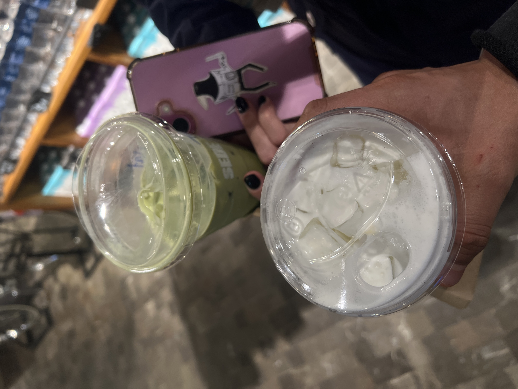 A high-angle shot shows two beverages in clear plastic cups held by two different people. The cup on the left contains a green liquid, possibly matcha, and the cup on the right contains a white liquid with ice cubes. The person holding the cup on the left has a pink smartphone with a sticker of an astronaut on the back. The scene appears to be indoors, with a blurred background suggesting a café or restaurant in Mexico City, Mexico.