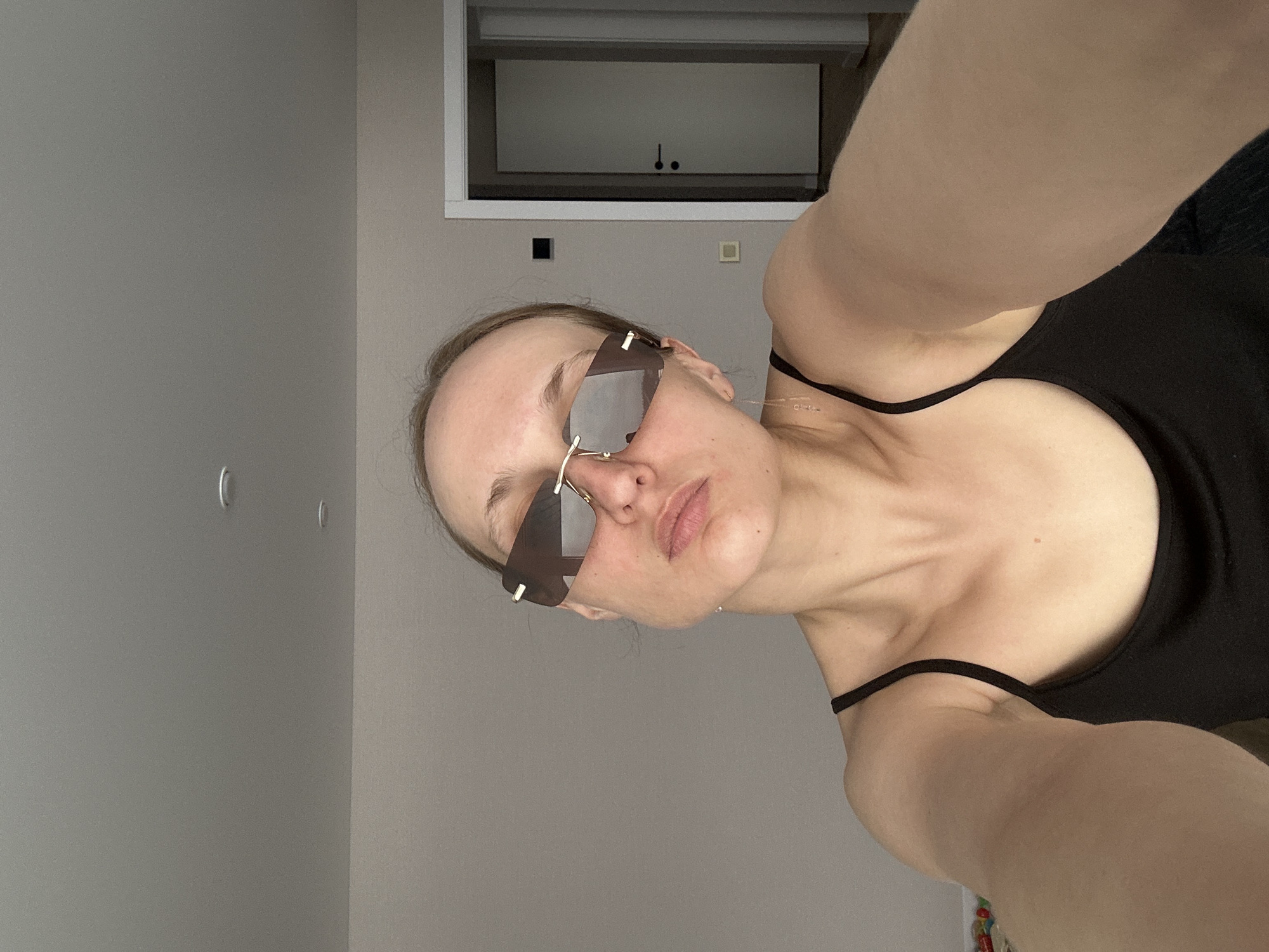 A woman in sunglasses and a black tank top is taking a selfie. The setting appears to be indoors, likely in a residence. A wall is visible, along with a window-like opening with a white frame and a dark interior. The image is taken at an angle, making it difficult to determine exact dimensions and spatial relationships within the scene. Given that location is in the city of Sepych, Russia, the style and setting could be consistent with a modern residential interior.
