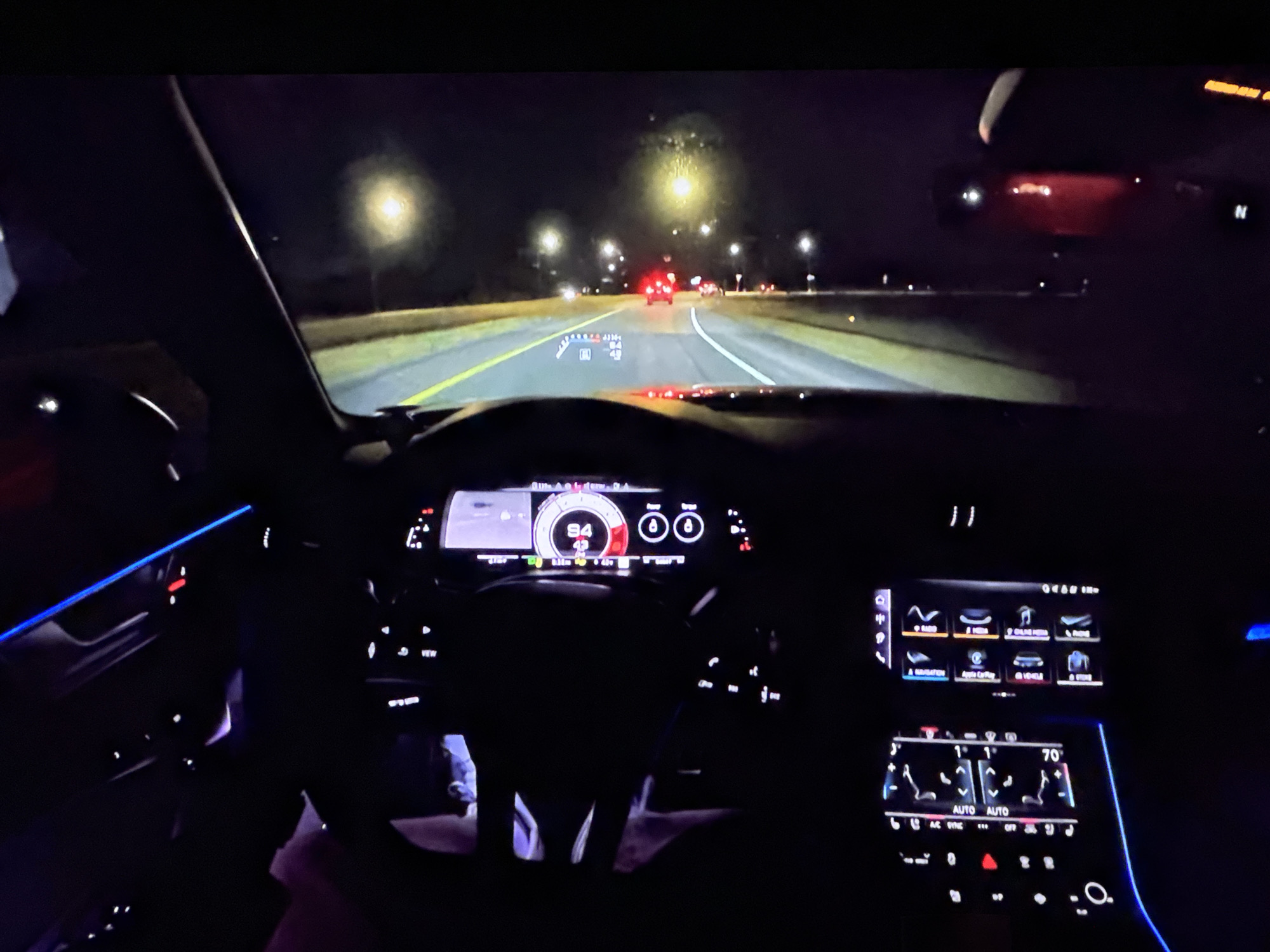The image is a first-person view from inside a car at night, driving on a highway. The dashboard displays illuminated screens showing speed and navigation, with ambient blue lighting visible on the left side of the car's interior. Streetlights line the road, and other vehicles are visible in the distance, with their taillights illuminated. The sky is dark, indicating it is nighttime. There are no people clearly visible in the shot, except for a hint of someone's legs in the driver's seat. The car appears to be traveling at a speed of 84 mph, as indicated on the dashboard. The environment suggests a typical highway driving scene at night. No specific details about the city of Bezden, Bulgaria, can be inferred from the image.