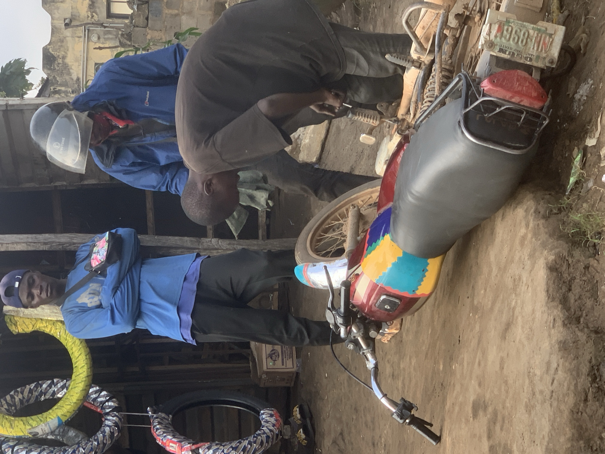 In Shagamu, Nigeria, at a roadside motorcycle repair stand, a dark-skinned man wearing a blue long-sleeved shirt and a dark baseball cap stands with his arms crossed, observing a motorcycle. Another man, also dark-skinned, wearing a dark t-shirt and dark pants, bends over the motorcycle, appearing to work on its engine. He has a dark helmet with a clear visor pushed up on his head. The motorcycle is lying on its side, its colorful seat visible. Behind the men, stacked tires hang from a makeshift wooden structure. The ground is dirt and debris.