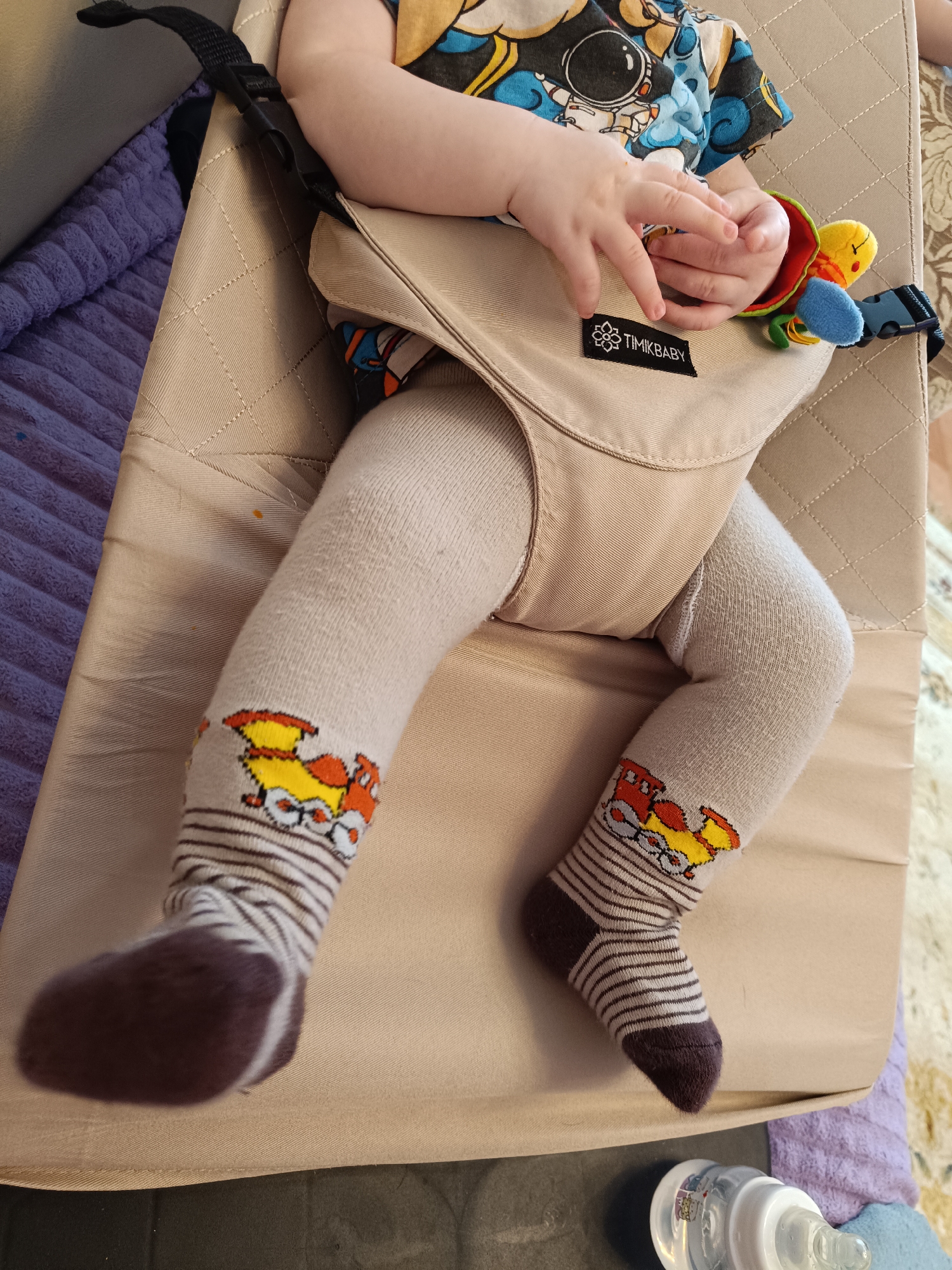 A baby is lying in a beige TIMIKBABY bouncer seat. The baby is wearing a t-shirt with an astronaut print and light beige tights with cartoon train socks. The baby is holding a small colorful toy attached to the bouncer. A baby bottle is visible near the bottom of the frame. The background includes a purple blanket and a patterned rug. There is no indication of the location being Tobolsk, Russia, or any specific city.