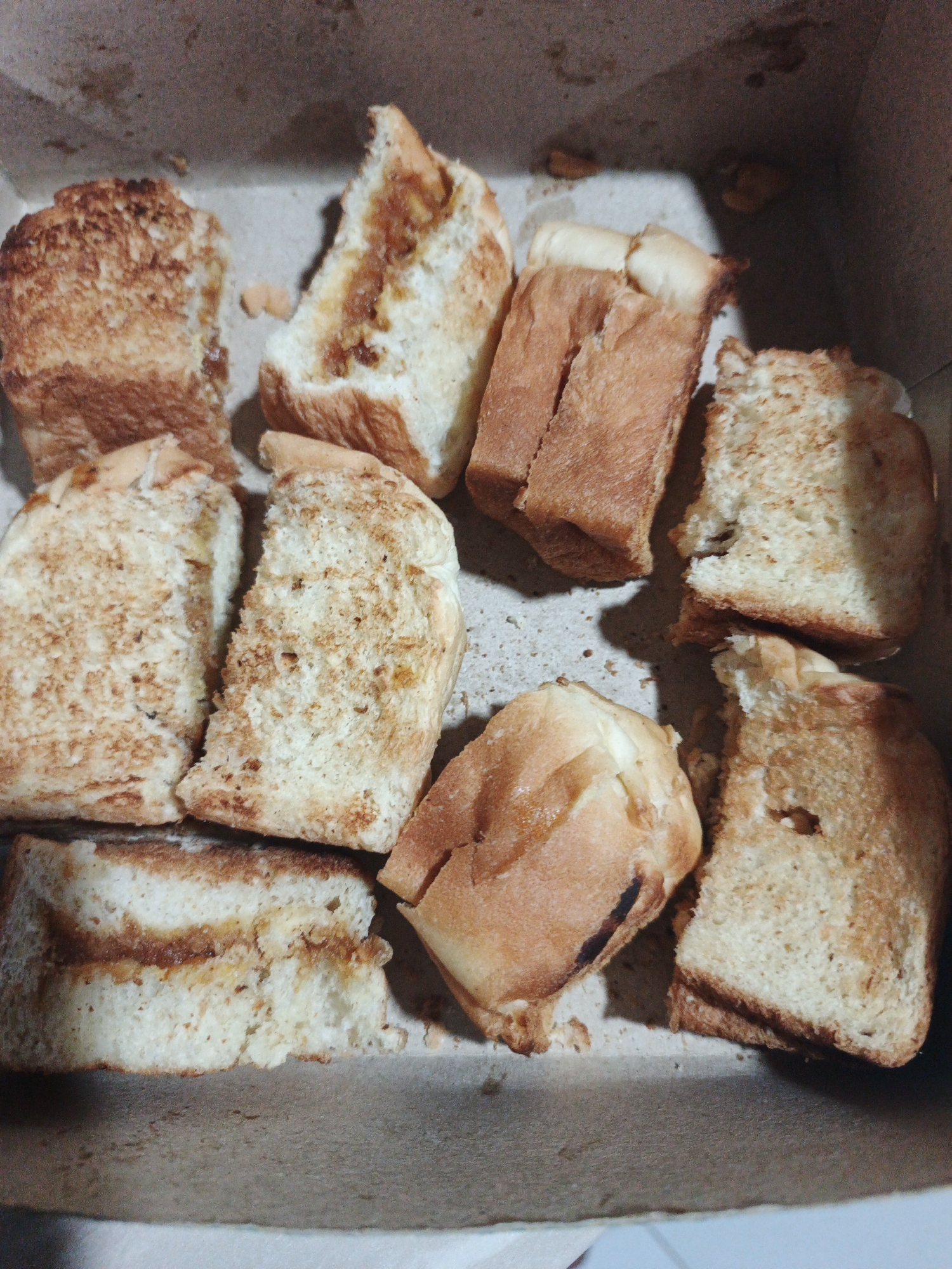 A close-up overhead shot shows a collection of toasted bread slices in a brown cardboard box. The bread is golden brown and some pieces appear to be lightly buttered or spread with jam. The slices are of various shapes and sizes, some are whole, while others are broken or torn. The box appears to be a simple take-out container. The lighting is dim, and the focus is on the bread. The scene is devoid of people or any clear indication of location beyond the food item itself. There are no discernible elements that would place this image specifically in Kelapapati, Indonesia.