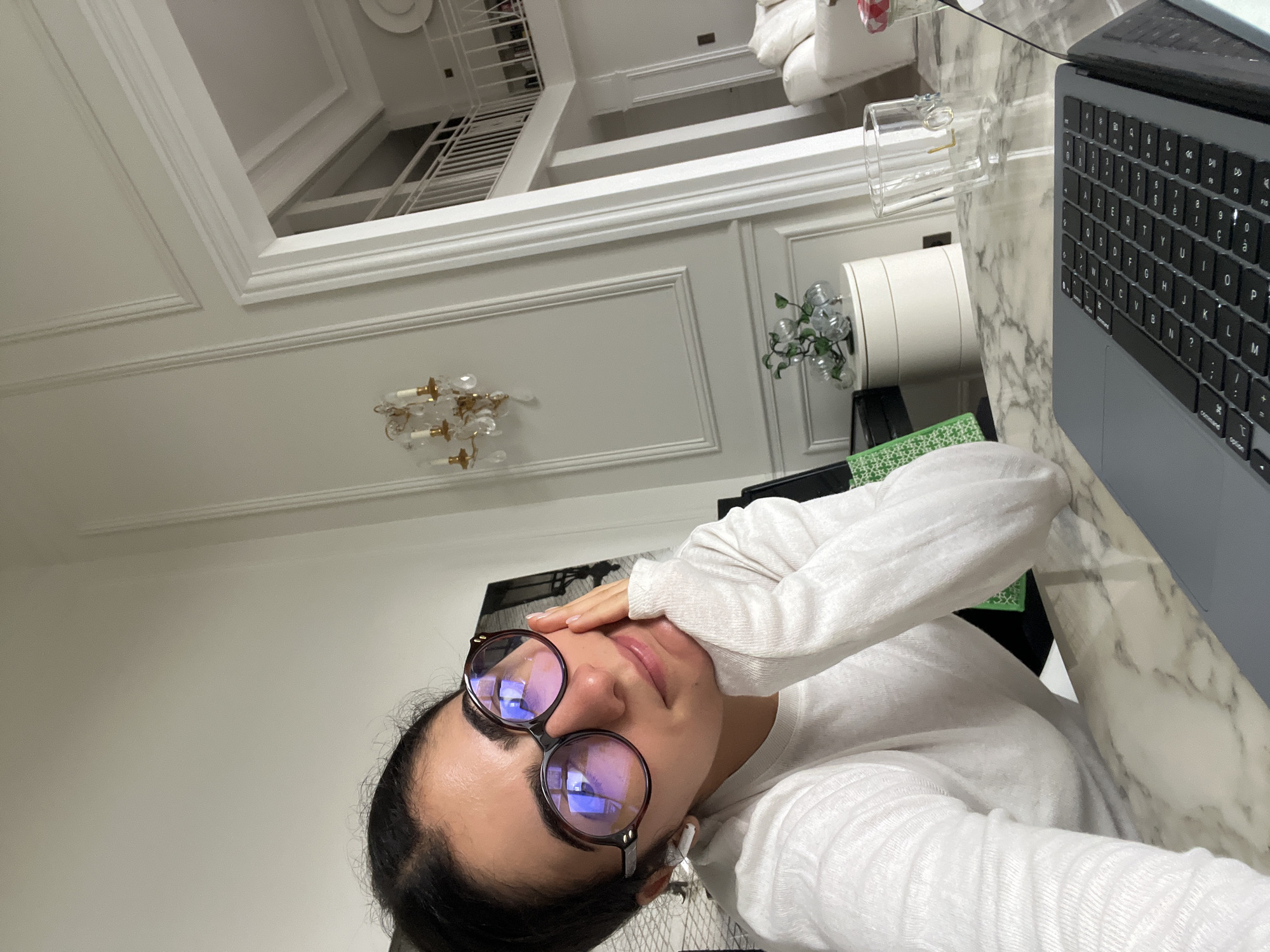 A woman wearing glasses is sitting at a desk with a laptop. The desk has a marble top, and there is a glass of water and a small plant on it. The room has white walls and a chandelier. There are stairs in the background. The location is likely a home or office in a city with ornate architectural details, possibly Paris, France.