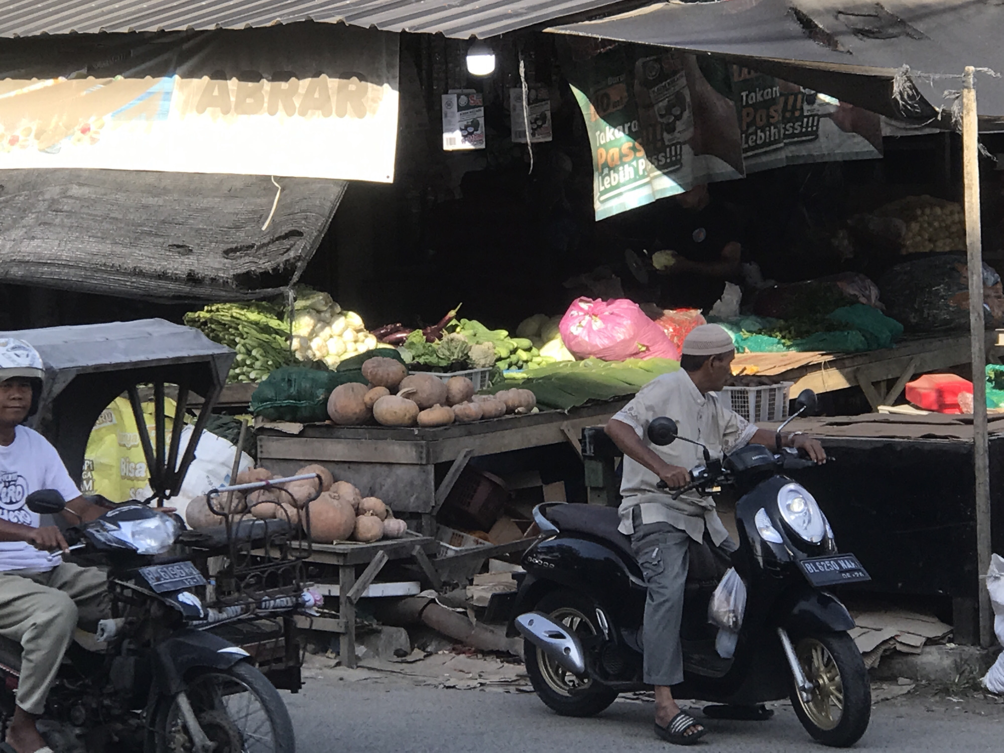 This image depicts a roadside market scene in Lhokseumawe, Indonesia. Two men are seen on motorbikes, one seated on a black scooter, the other on a black motorcycle with a front basket. The market stall behind them is laden with produce, including pumpkins, squashes, and various green vegetables. Banners and advertisements are hanging above the stall, with signs in Indonesian like "Takara Pass Lebih Pass!!!" and "Takan Pos!!!" A sign with the name "ABRAR" is also visible. The scene is bustling, with the market offering a variety of produce for sale.