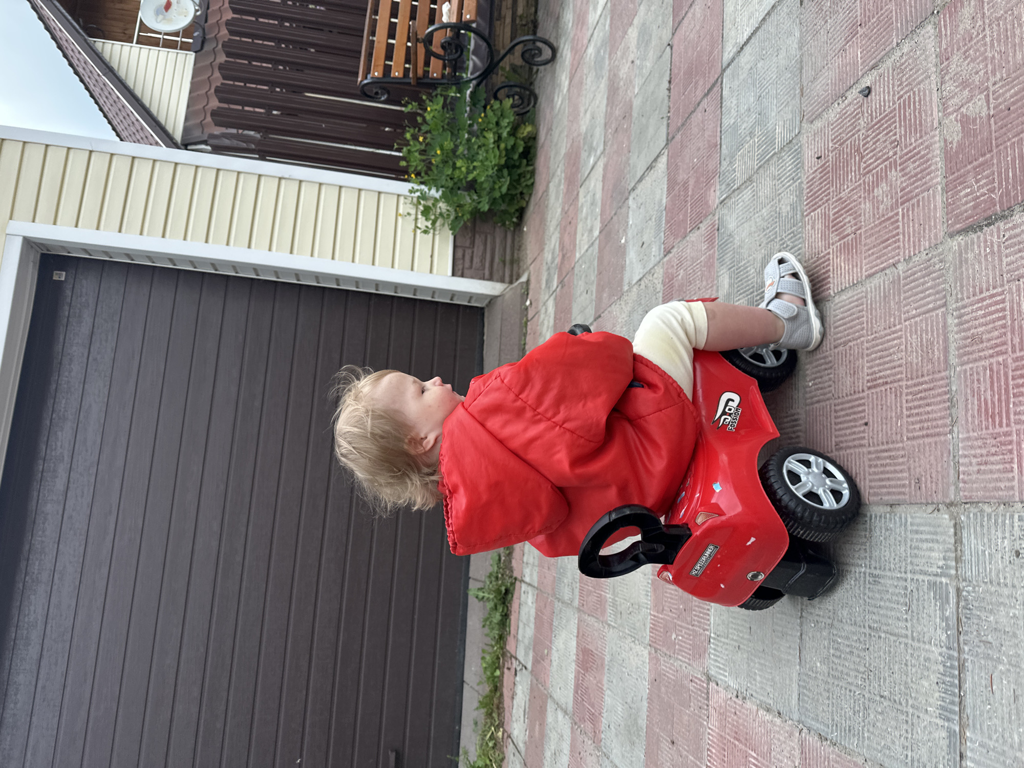 A young child is sitting on a red toy car on a paved surface consisting of alternating red and gray square tiles. The child is wearing a red jacket and light-colored pants, and a gray sandal. The child is facing to the left. Behind the child is a dark gray garage door. To the left of the garage door is a beige wall with white trim. Further back is a section of a brown roof. Above the beige wall is a wrought iron plant stand holding a green plant. The image appears to be taken outdoors. There is no information in the image to suggest it is in Nizhny Novgorod, Russia.