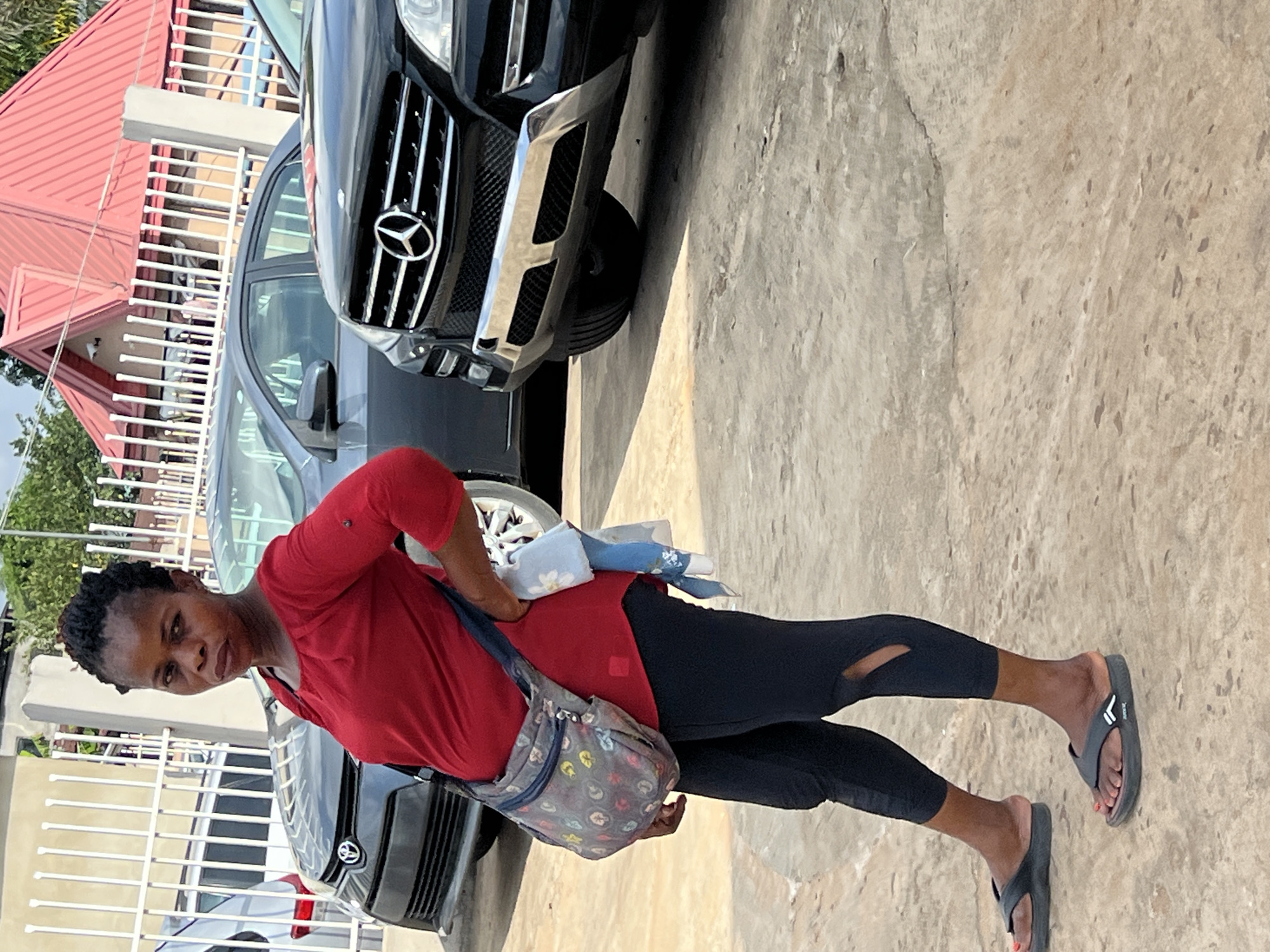The image, originally rotated, depicts a woman standing outdoors in bright daylight. She faces the camera with a neutral expression, wearing a red short-sleeved top and black leggings with cut-outs at the knees. Her feet are clad in dark-colored flip-flops, revealing red-painted toenails. She has short, dark hair, possibly braided. A grey, patterned cross-body bag is slung over her left shoulder, and she holds some folded light-colored items in her right hand near her waist.
The setting appears to be a paved area with a light-colored, textured concrete ground. In the background, a white metal fence runs horizontally, behind which a building with a prominent red pitched roof is visible. Green foliage can be seen further in the distance.
Two vehicles are present. A black Mercedes-Benz SUV, clearly identifiable by its distinctive star emblem on the grille, is parked in the mid-ground on the right. Another dark-colored car is partially visible behind the woman and to the left of the Mercedes.
The scene is brightly lit, suggesting clear weather conditions during the daytime. There are no overt activities taking place, and the woman appears to be posing for the photograph. Visible text includes the Mercedes-Benz logo, and faint branding on the flip-flops, which seems to be "XONOX" on one and "JOBUXX" on the other. The environment is consistent with an urban or residential area, aligning with the provided location context of Onitsha, Nigeria.