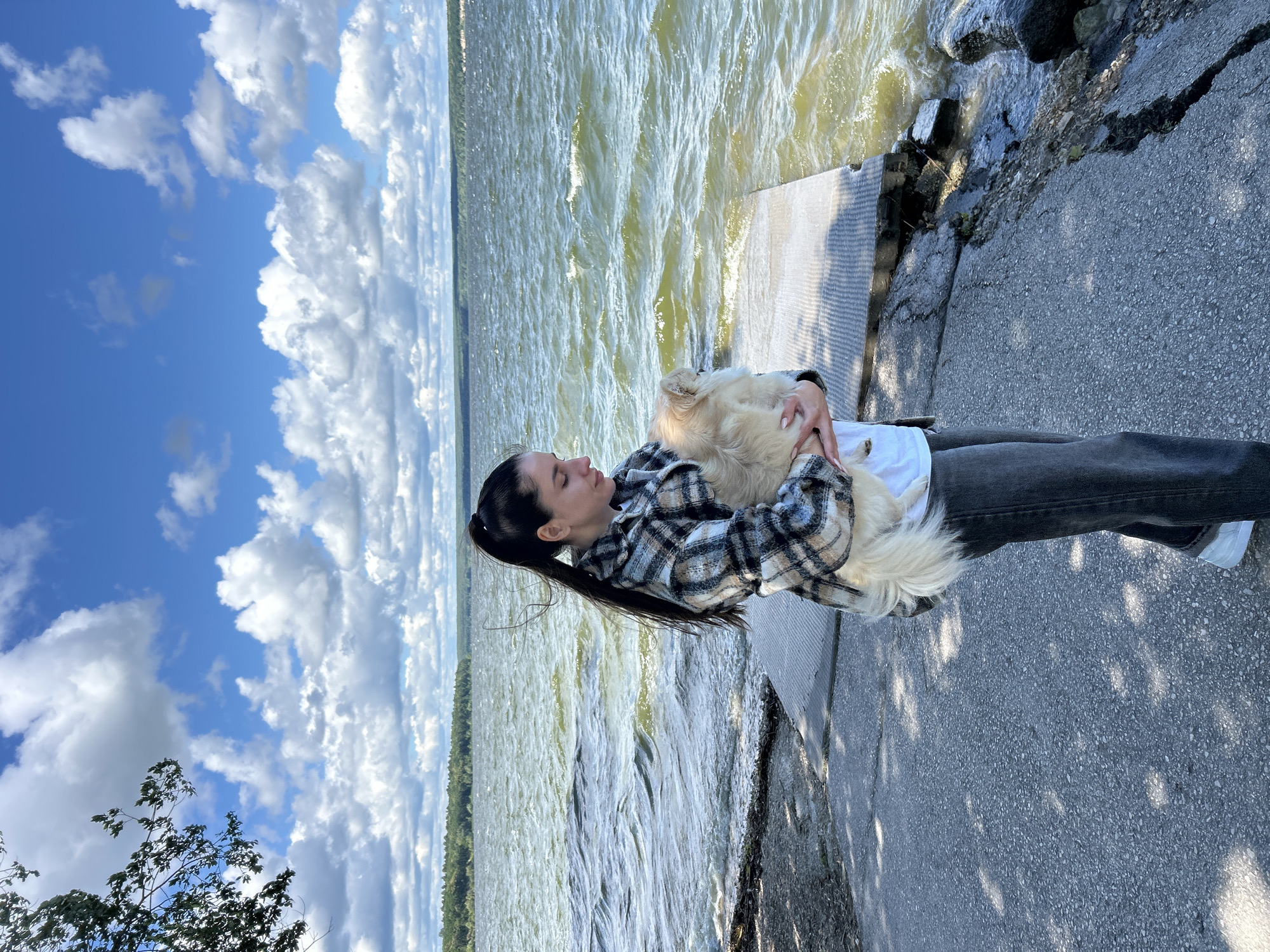 A young woman with dark hair pulled back in a ponytail is holding a light-colored fluffy dog. She is wearing a plaid jacket and dark jeans. They are positioned on a concrete walkway next to a body of water, which appears to be a lake or river, with choppy waves. The background shows a distant tree line and a bright blue sky filled with dramatic, puffy white clouds. The overall lighting suggests it is daytime, likely late morning or afternoon, with the sun casting shadows on the ground. The woman's eyes are closed, and she appears relaxed, cradling the dog close to her. The setting feels serene and natural, with the emphasis on the bond between the woman and her pet against the backdrop of a vast sky and water. There are no visible signs of text in the image.