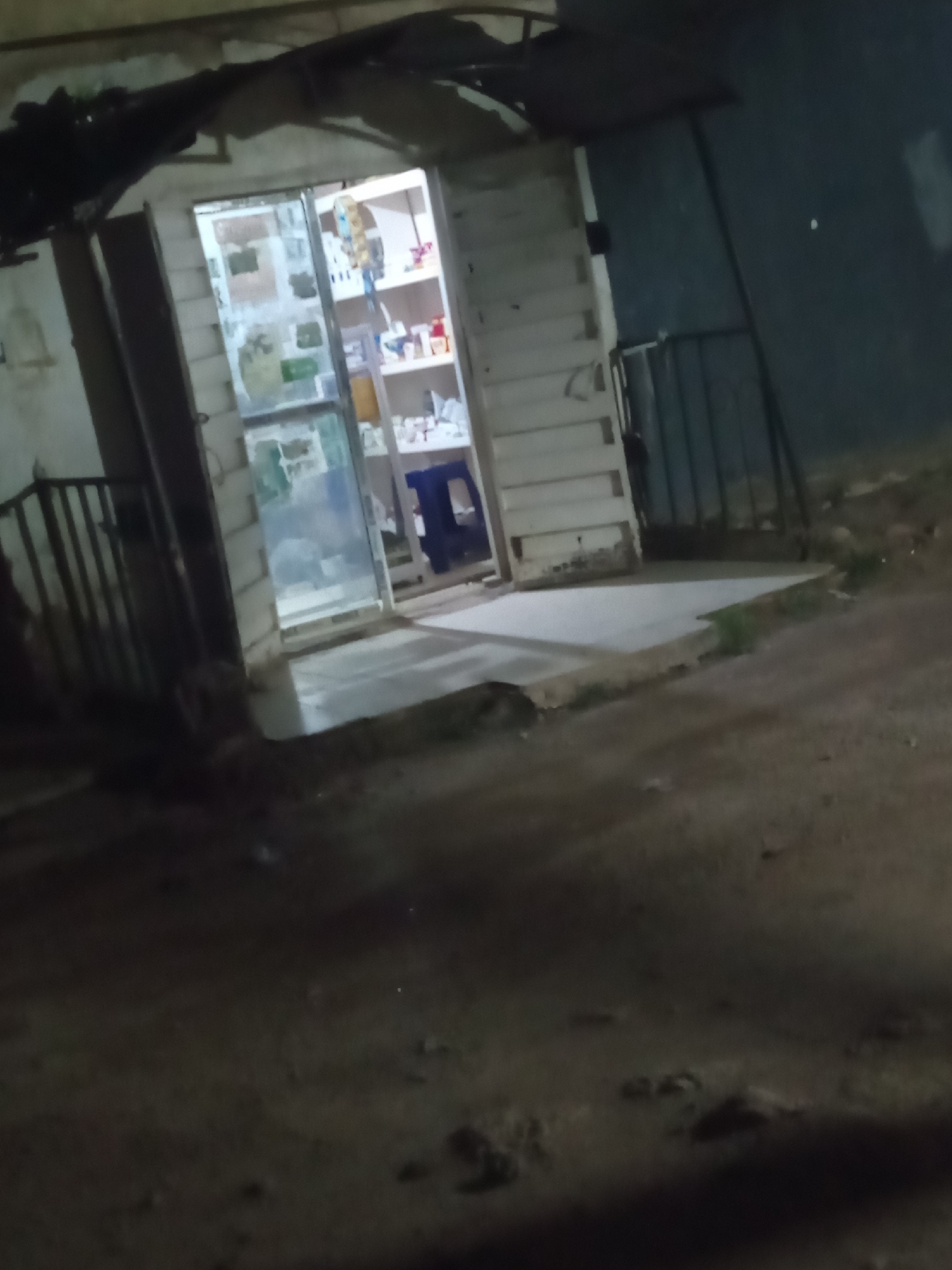 The image shows a small storefront at night, likely a local shop or pharmacy, with a partially open glass door revealing shelves stocked with goods. The structure appears to be made of wooden planks and corrugated metal, with a makeshift awning. Metal fencing is visible alongside the building. The surrounding environment is dark and appears to be a street or path. The location is described as Jos, Nigeria. No people are visible in the image.
