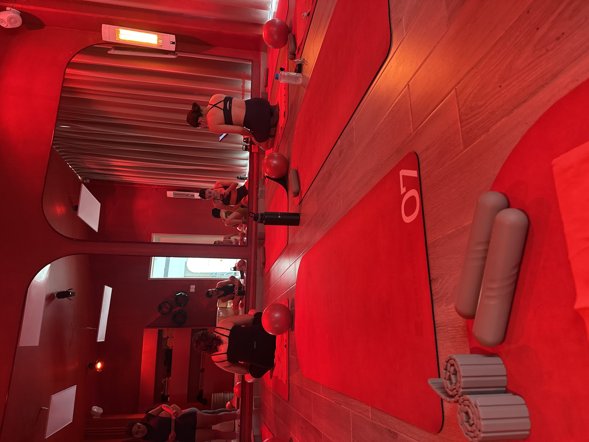 The image shows a group of people in a fitness studio with red lighting. In the foreground, there are two rolled-up mats and two cylindrical foam rollers on a red yoga mat. Further back, several other red yoga mats are laid out, some with red exercise balls on them. A woman in a black tank top and shorts is standing to the left, facing away from the camera. In the middle ground, a woman in a black tank top and leggings is sitting on a mat in a cross-legged position, looking at her phone. To her right, another woman in a black sports bra and leggings is kneeling on a mat, also looking at her phone. In the background, several other individuals are visible sitting or kneeling on mats, some of whom are reflected in a large mirror that covers much of the wall. The room has a wooden floor and a red ceiling with some recessed lighting fixtures. A white infrared heater is mounted on the wall on the right. There is text visible on the mirror that reads "@redmethodmx". This type of studio is characteristic of a boutique fitness chain, and without additional information, the specific location in Puebla, Mexico cannot be determined.