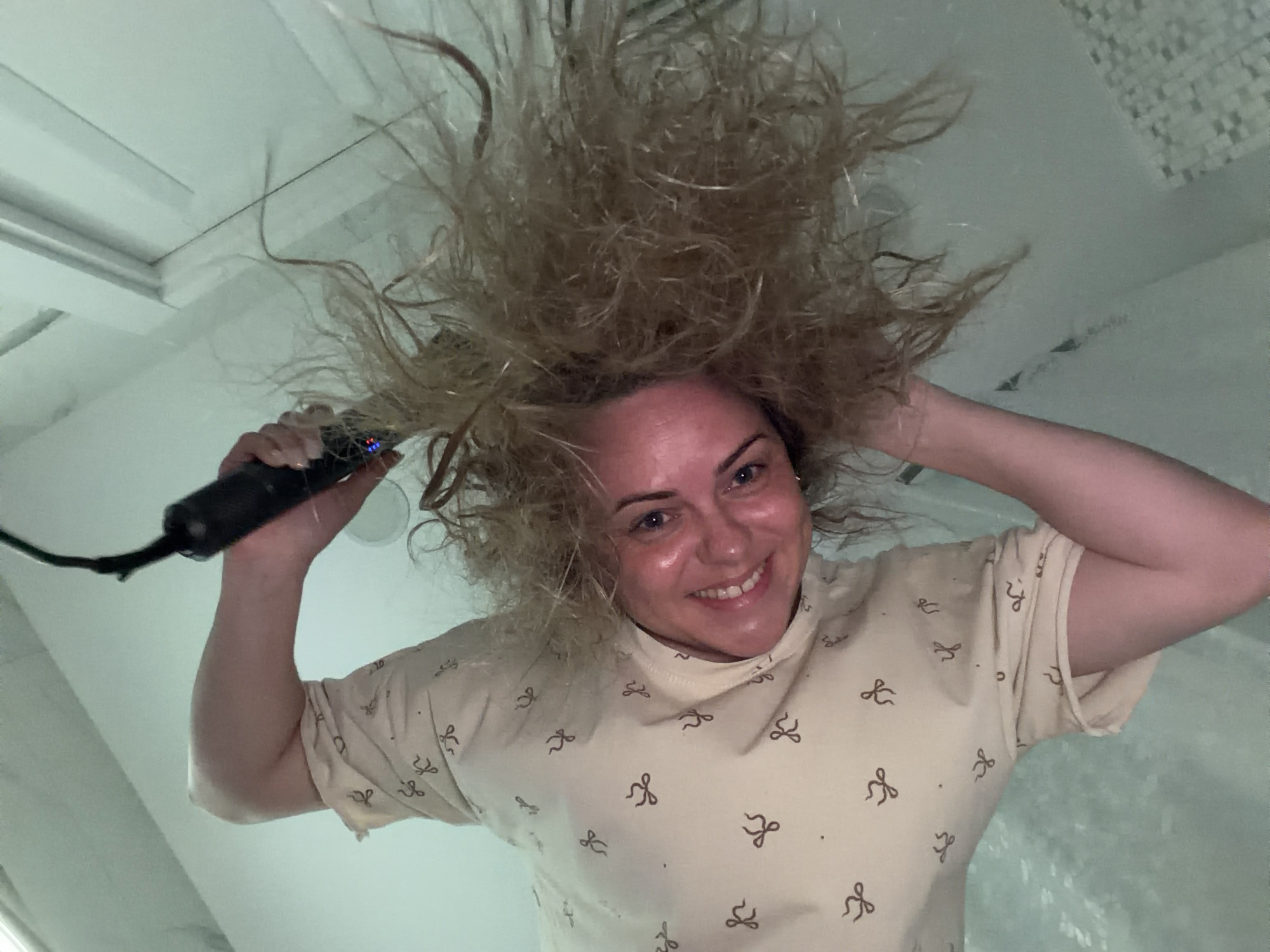 Captured in Votkinsk, Russia, this image features a woman from a low-angle perspective, looking directly down into the camera with a wide, joyful smile. Her blonde hair is spectacularly disheveled and voluminous, standing almost upright, as if she has just been blow-drying it with an electric charge effect.
In her left hand, she holds a black hair dryer or styling tool, pointed towards her head, reinforcing the action of styling her wild hair. Her right hand is raised, with fingers spread, contributing to the dynamic and playful pose. She wears a light-colored, possibly cream or beige, short-sleeved t-shirt patterned with small, dark bow-tie shapes.
The background is a bright, minimalistic indoor setting, predominantly white or light grey, with subtle architectural lines that suggest a room with a sloped ceiling or a corner. Some areas in the background show what appears to be small, light-colored tiles, commonly found in bathrooms. The overall lighting is bright and uniform, creating a clean, airy feel. The woman's expression is one of amusement and happiness, highlighting the lighthearted nature of the scene. No visible text is present in the image.