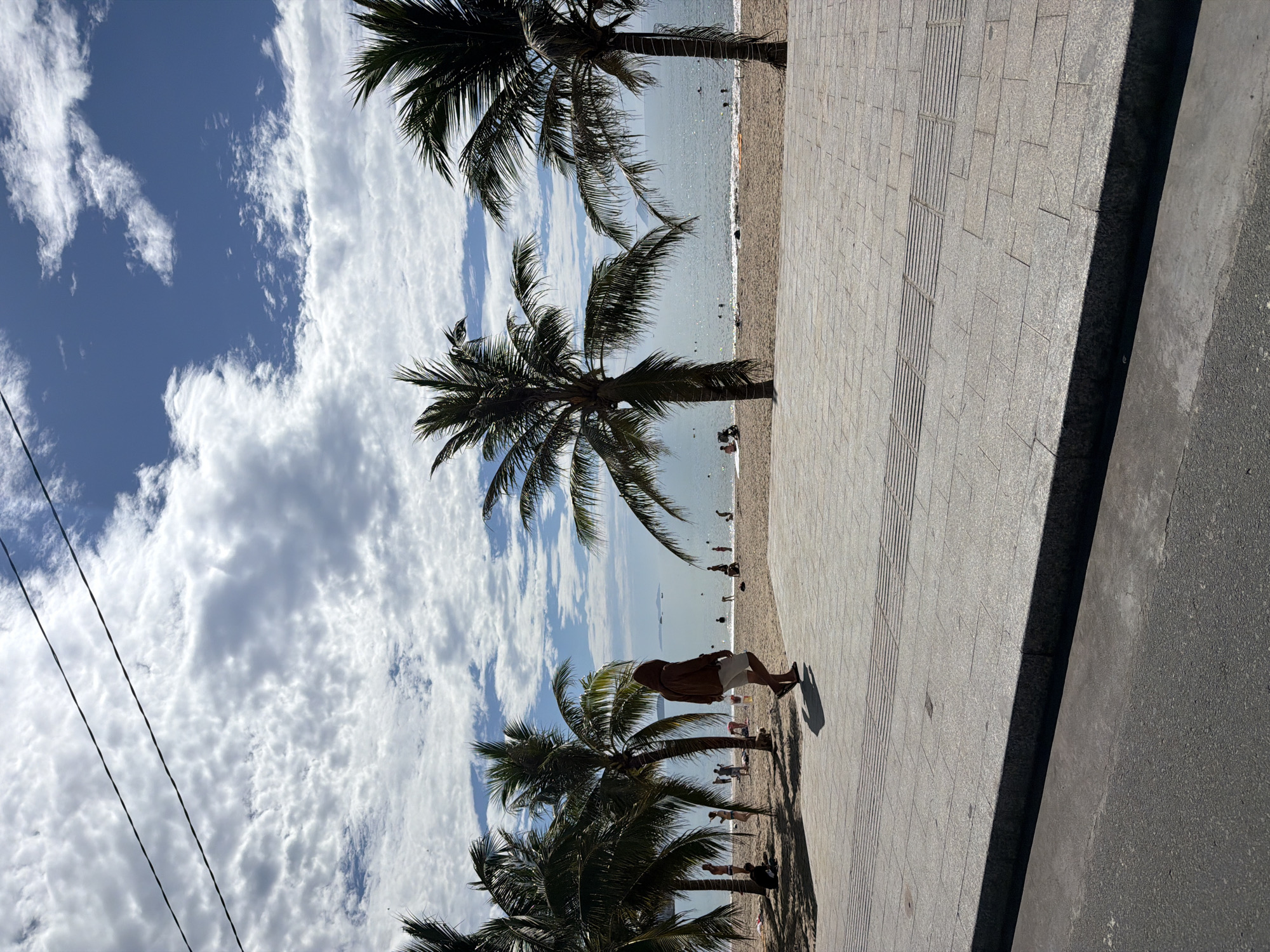 The image captures a vibrant, sunlit coastal scene in Nha Trang Ward, Vietnam, centered around a beach, the sea, and a paved promenade. The viewpoint is from the promenade, looking towards the water.
In the foreground, a wide, light-grey paved walkway, constructed from rectangular tiles, stretches across the left side of the frame. A strip of tactile paving is integrated into this surface. Adjacent to it, on the far left, is a darker asphalt road. A lone figure is walking along the promenade, seen from behind, wearing light shorts and a dark top, with their shadow extending before them.
A series of tall palm trees with abundant green fronds lines the edge of the promenade, casting shadows and partially obscuring the sandy beach and sea beyond. The sandy beach, populated by several people enjoying the day, extends towards the sparkling blue-green ocean. Numerous individuals are visible on the sand and swimming or wading in the clear water. In the distance, the horizon separates the sea from a bright blue sky, with faint landmasses or islands visible.
The sky dominates the upper part of the image, showcasing a striking contrast of deep blue patches interspersed with large, fluffy white cumulus and altocumulus clouds. In the upper left corner, two dark power lines are visible, stretching diagonally across the cloudy sky. The bright light and distinct shadows indicate it is daytime, likely mid-day, under clear and sunny weather conditions. There is no visible text in the image.