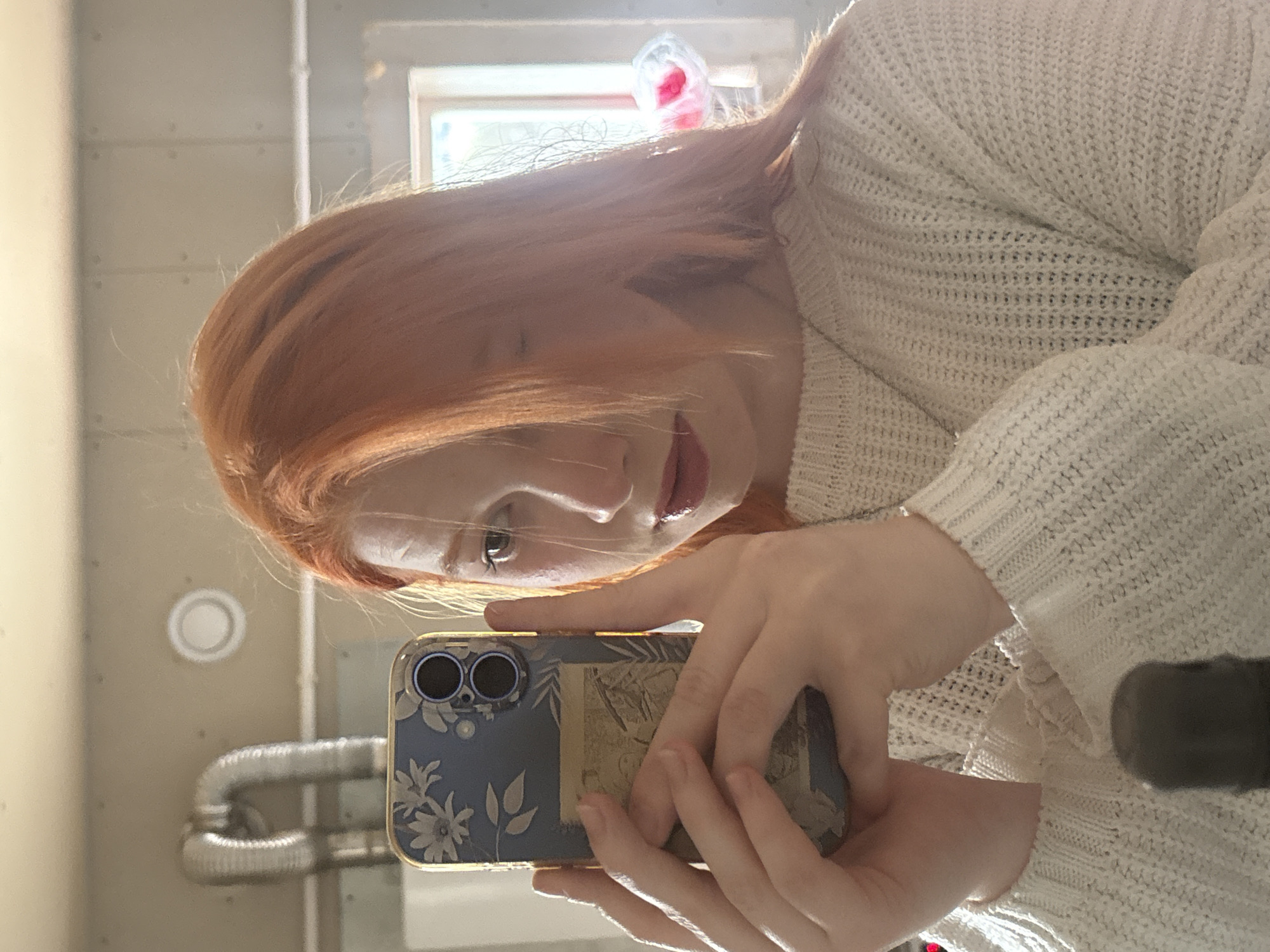A mirror selfie features a person with long, reddish-brown hair and fair skin, wearing a light-colored, possibly cream or white, knitted sweater. The person's face is partially visible, showing one eye and glossy lips with a neutral expression, as they look down towards the smartphone they are holding.
The smartphone has a clear case with a blue background and a white floral pattern. Two dark camera lenses are visible on the back of the phone. Through the clear case, a rectangular insert or sticker depicting what appears to be an antique-style map or illustration is visible.
The background is a reflection of an indoor setting. Light-colored walls with horizontal seams or panels are visible. On the left, there's a circular white vent or light fixture. Below it, silver, flexible ductwork runs horizontally. A window in the upper right background allows natural light to enter, and a crumpled red object is visible near the window frame. The overall lighting suggests it is daytime. No visible text is discernible on the phone or in the background.