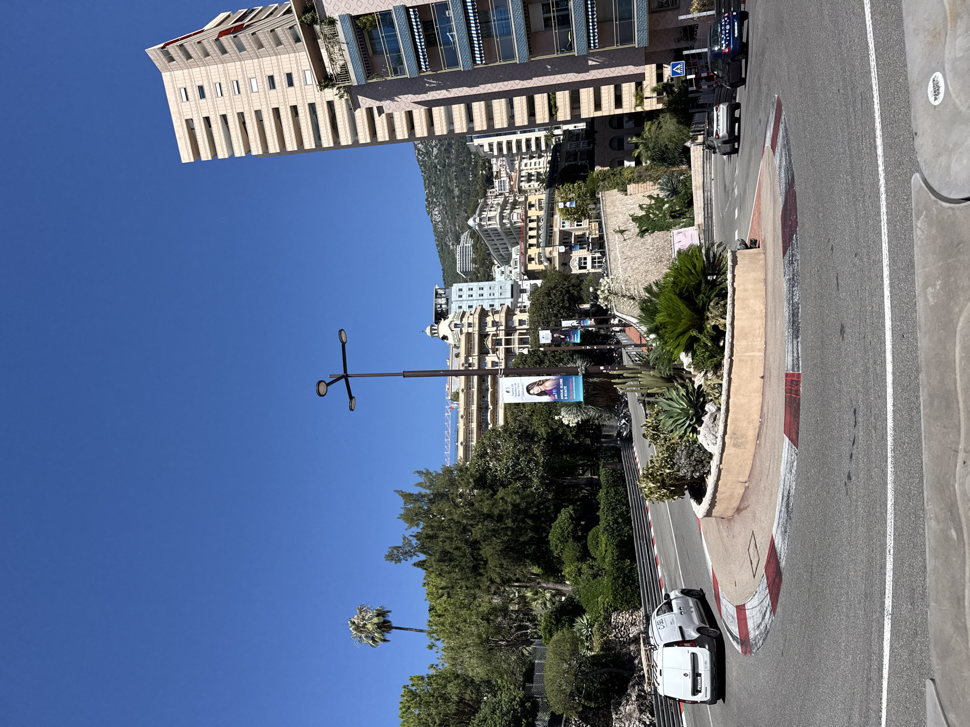 In the city of Monaco, Monaco, a clear blue sky presides over a race circuit. The foreground features a roundabout with a planter filled with cacti and palm-like plants. A white utility van with the text "CI-SEL" on its side is turning left on the circuit. Beyond the roundabout, a light grey SUV and a blue car are visible further down the road. Tall, modern buildings with balconies and awnings dominate the background, with a larger, beige building on the right featuring many windows. Lush green trees and smaller plants are interspersed throughout the scene, creating a vibrant urban landscape. A banner advertising "AM 10: AMOUR, GLOIRE & BEAUTE" hangs from a lamppost on the right side of the circuit. The road has red and white curbs, indicating a race track.