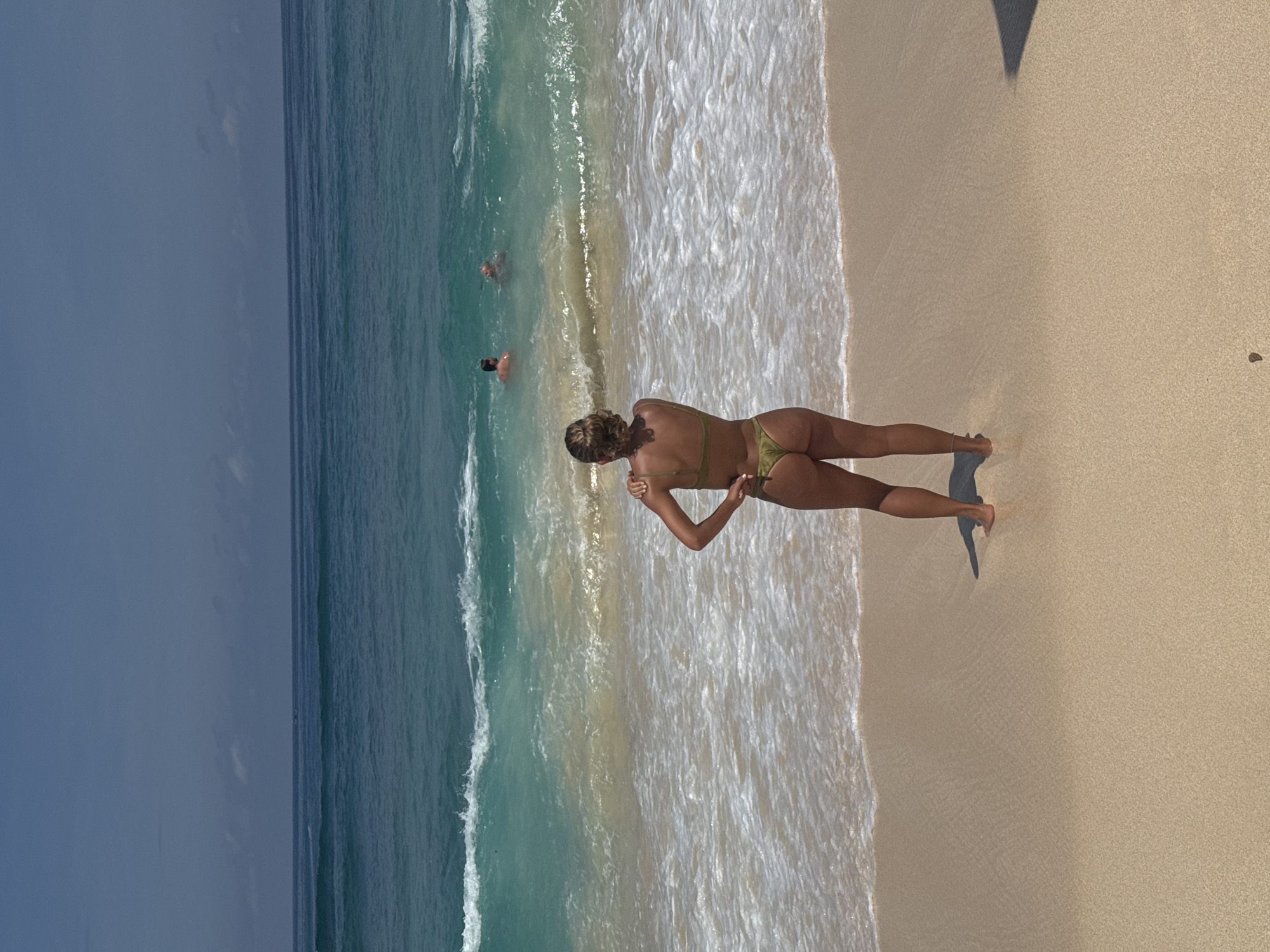 The image shows a woman standing on a sandy beach next to the ocean in Kuta Selatan, Indonesia. The woman is wearing a green bikini and is seen from behind. Two people are visible swimming in the water. The sky is overcast. The ocean is turquoise near the shore, becoming darker blue further out. Waves are crashing onto the beach. The sand is light-colored. A shadow is cast on the sand near the woman.