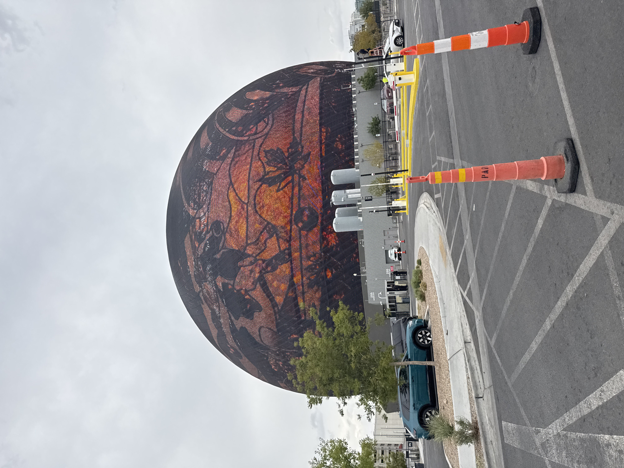 The image shows the exterior of the Sphere, a large geodesic dome entertainment venue located in Paradise, Nevada, United States of America. The dome is covered in a vibrant, illuminated display depicting abstract shapes and patterns in shades of orange, yellow, and brown. In the foreground, a dark teal SUV is parked on the left, next to a small patch of green bushes and gravel. Several traffic cones, orange and white with black bases, are placed on the asphalt. A white car is visible on the right side of the image, near a yellow and white barrier gate. The sky is overcast, with a pale grey cloud cover. A few trees with green foliage are scattered around the scene. The background includes a low-rise building with industrial-looking elements and some taller structures in the distance.