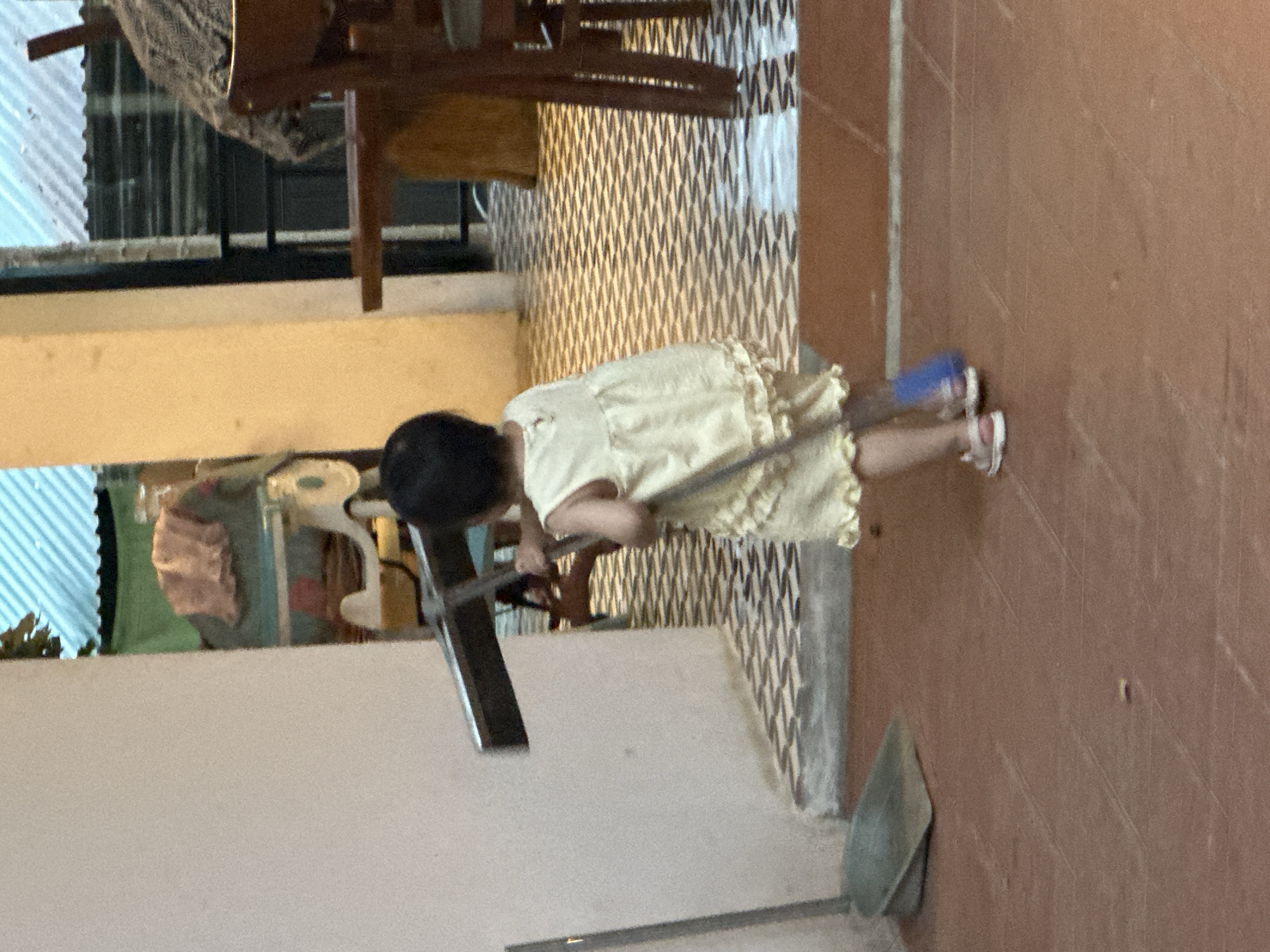 A young child, wearing a light yellow ruffled dress and white sandals, is standing on a red tiled floor, holding a broom and dustpan. The child is facing away from the camera and appears to be sweeping. The broom is made of wood and has blue bristles. The dustpan is made of metal and is green in color. The floor is tiled with a pattern of brown and white squares. In the background, there is a wall with a white textured finish, and a doorway leading into another room. The other room has a checkered floor and a wooden table with chairs. The image appears to have been taken in Cổ Lũng Commune, Vietnam, given the presence of a tropical roof structure and the general architecture.