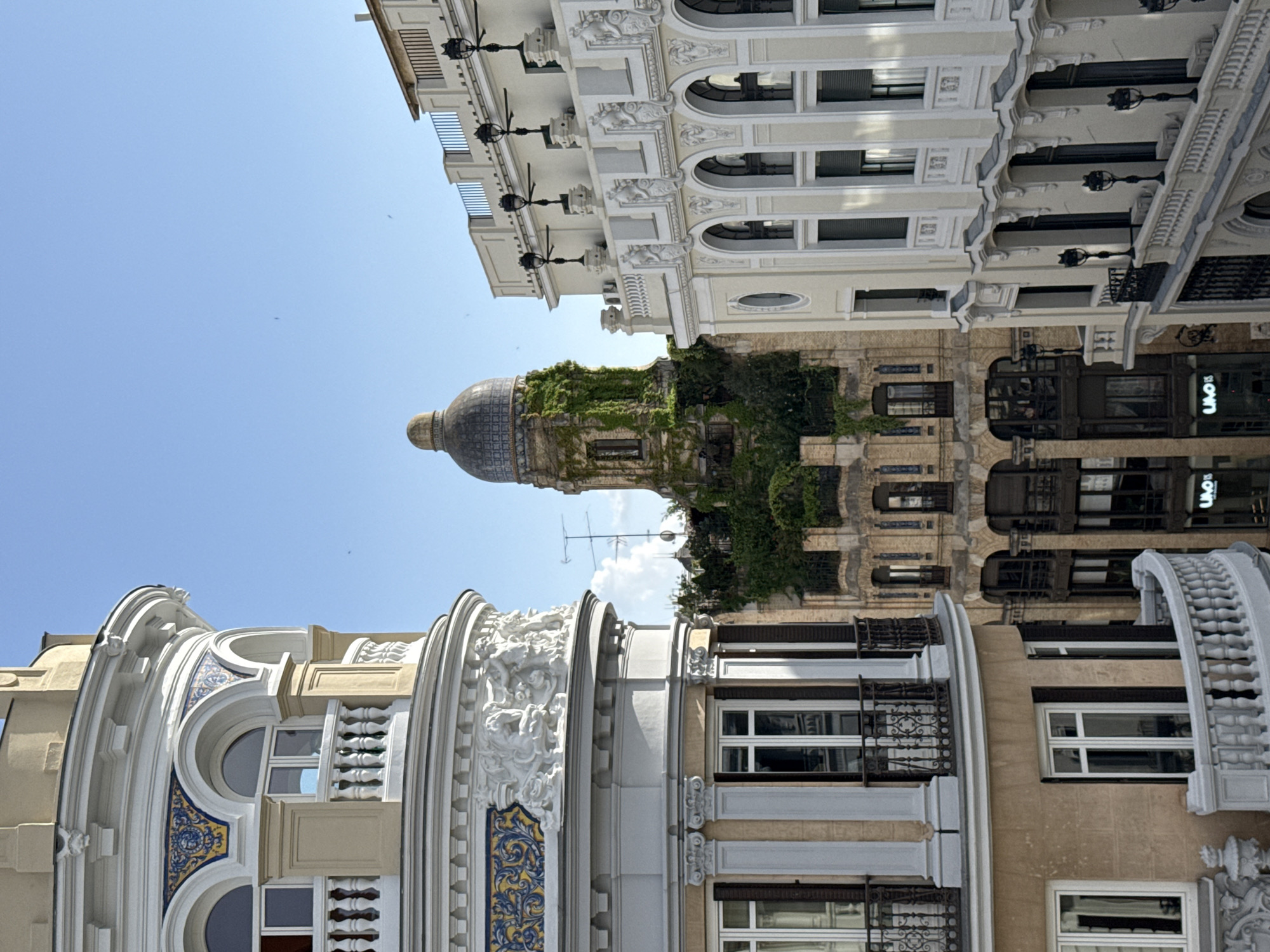 The image captures an upward-looking perspective of ornate buildings in an urban setting, identified as Madrid, Spain, under a clear blue sky. The scene is brightly lit by natural daylight, suggesting midday or early afternoon.
In the foreground and to the left, a building features a curving, light yellow-ochre facade adorned with intricate white stucco decorations, including balustrades and sculptural elements. Notable details are sections of vibrant blue and yellow patterned tiles (azulejos) around its round and rectangular windows.
The dominant structure in the midground, to the right, is a grand multi-story building with a light beige and white facade. It boasts numerous windows, some arched, and ornate balconies with black wrought-iron railings. Decorative white moldings, cornices, and pilasters enhance its classical or Art Nouveau aesthetic. Several black, decorative street lamps extend from its facade.
In the background, positioned centrally, a dome-shaped structure, possibly a cupola, is heavily covered in lush green ivy, giving it a weathered, organic appearance. Below the dome, more of its underlying building is visible, also partly obscured by greenery. To the far right, a darker building facade is partially visible, featuring a clear, illuminated white sign displaying the text "UAOIS."
The sky is a brilliant, cloudless blue. A faint weather vane or antenna is discernible against the sky above the ivy-covered dome. No people are present in the static architectural scene, which emphasizes the rich detailing and historical character of the city's buildings.