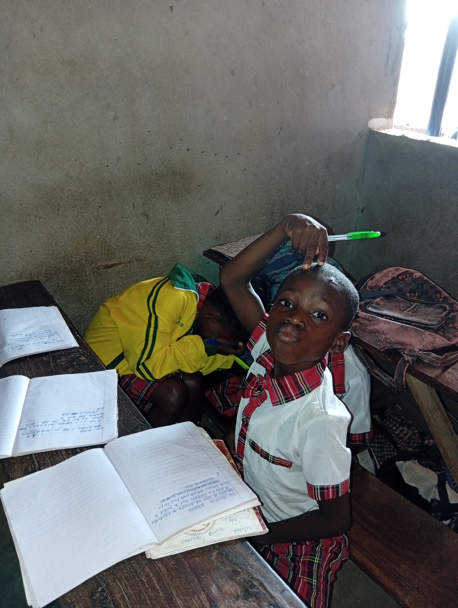 A young Black boy in Ukwu-Abwa, Nigeria, sits at a wooden desk in a classroom. He wears a white shirt with red and green plaid trim and matching plaid shorts. He holds a green pen over his head with his right hand and makes a pouting face at the camera. An open notebook with handwriting is on the desk in front of him. Another child, wearing a yellow and green track suit, is slumped over at the desk next to him. Another open notebook is on that desk. A backpack sits on the desk behind the first boy. The classroom has rough, undecorated walls. Sunlight enters from a window.