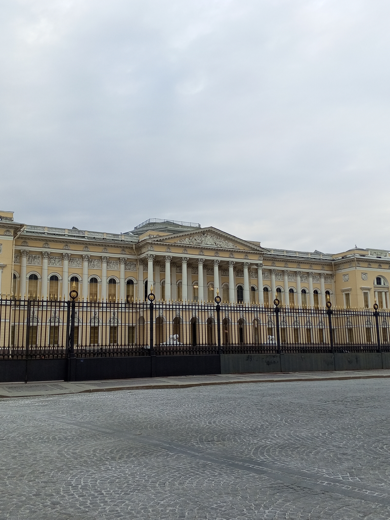 The image shows the facade of the Mikhailovsky Palace (also known as the Russian Museum) in Saint Petersburg, Russia. The palace is a pale yellow building with a long row of white columns in the center. A decorative frieze runs along the top of the column section. A black metal fence with gold ornamental details runs along the perimeter of the palace grounds, partially obscuring the building. The ground in front of the fence is paved with cobblestones. The sky is overcast with gray clouds. There are no people clearly visible in the image.